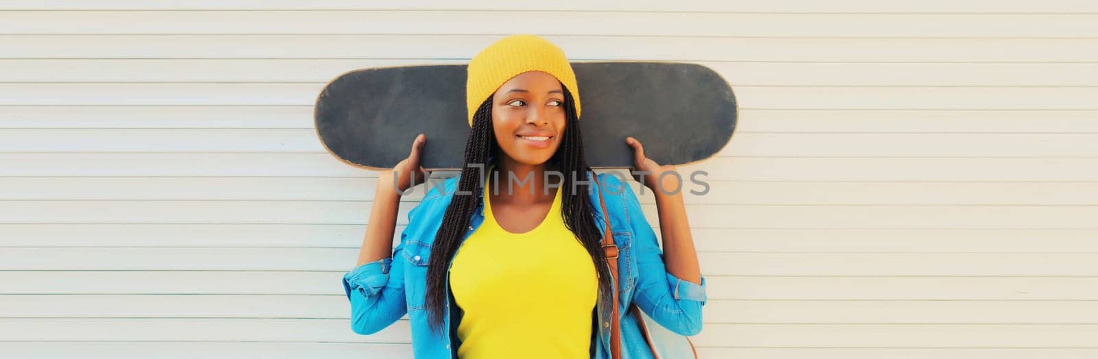 Happy smiling young african woman in colorful yellow hat posing with skateboard in the city on white wall background