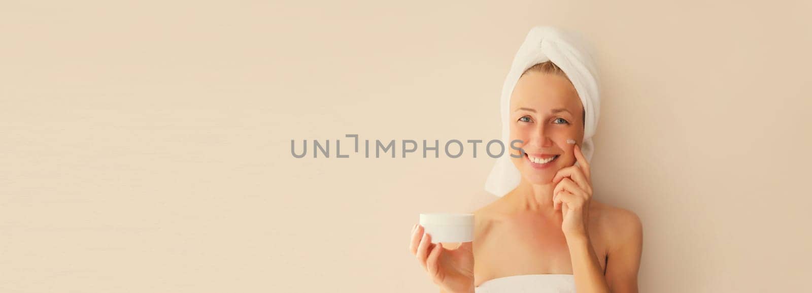 Natural beauty portrait of happy smiling caucasian middle-aged woman touches her clean skin applying face cream drying wet hair with white wrapped bath towel on her head after shower in morning