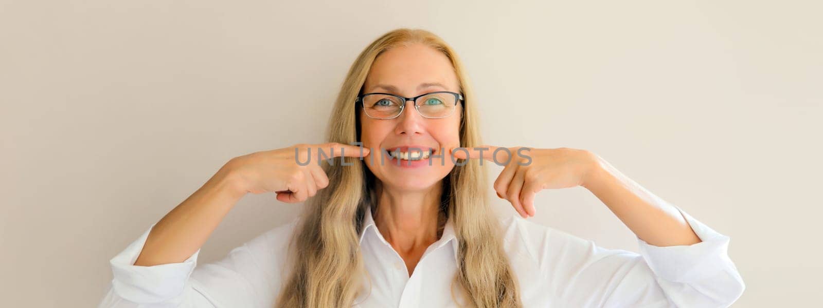 Portrait of happy smiling middle-aged woman points with fingers to her white clean teeth in eyeglasses on white studio background