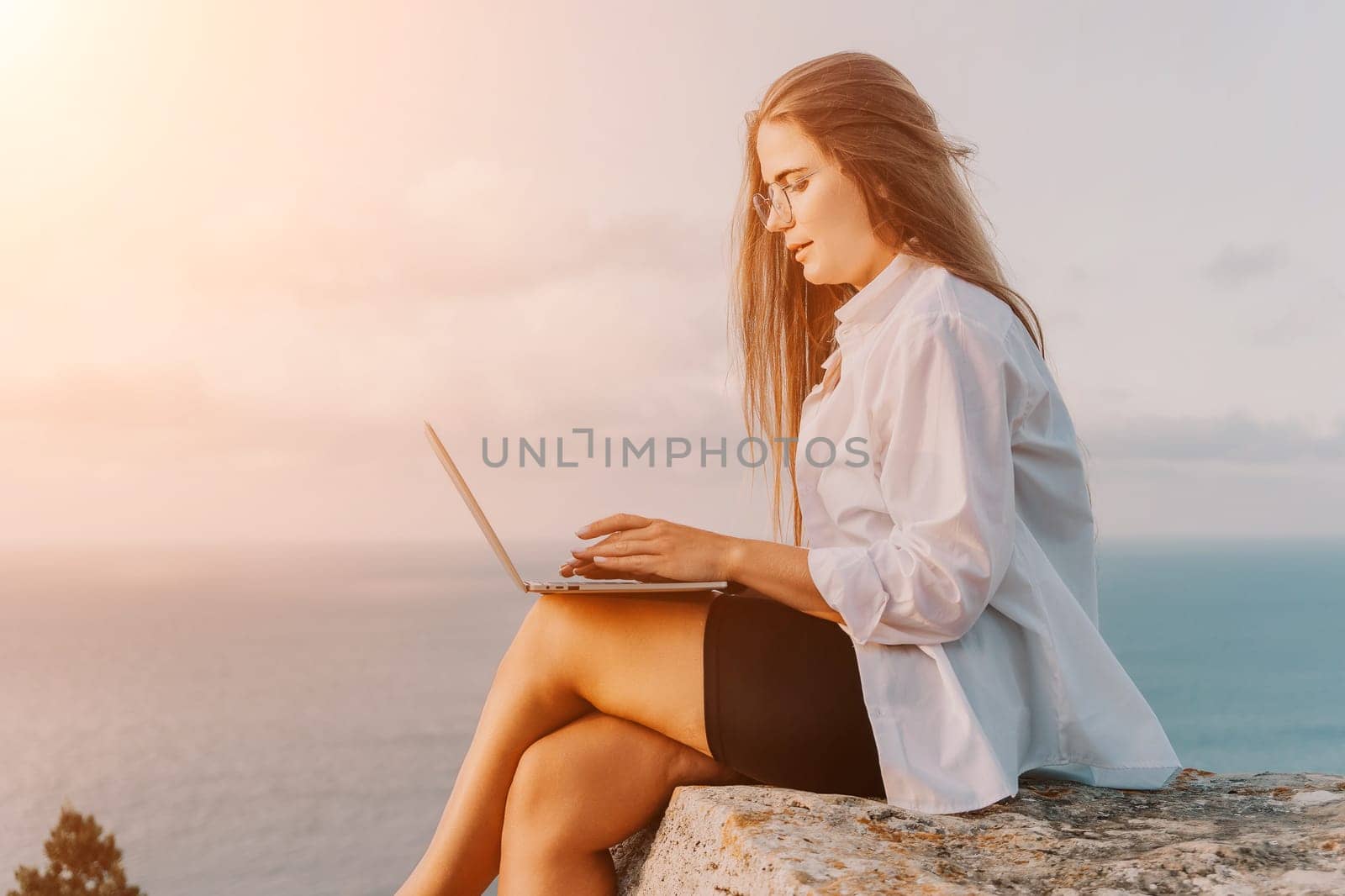 Digital nomad, woman in the hat, a business woman with a laptop sits on the rocks by the sea during sunset, makes a business transaction online from a distance. Freelance, remote work on vacation.