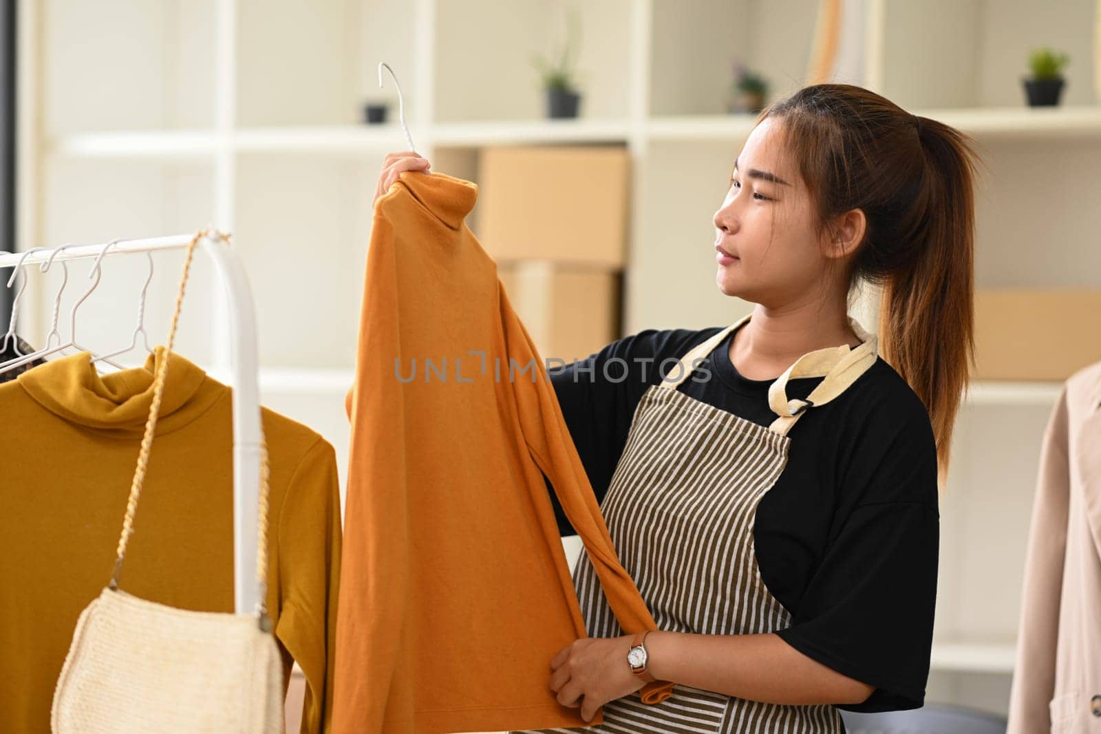 Smiling young female clothing store owner in apron arranging clothes on rack.