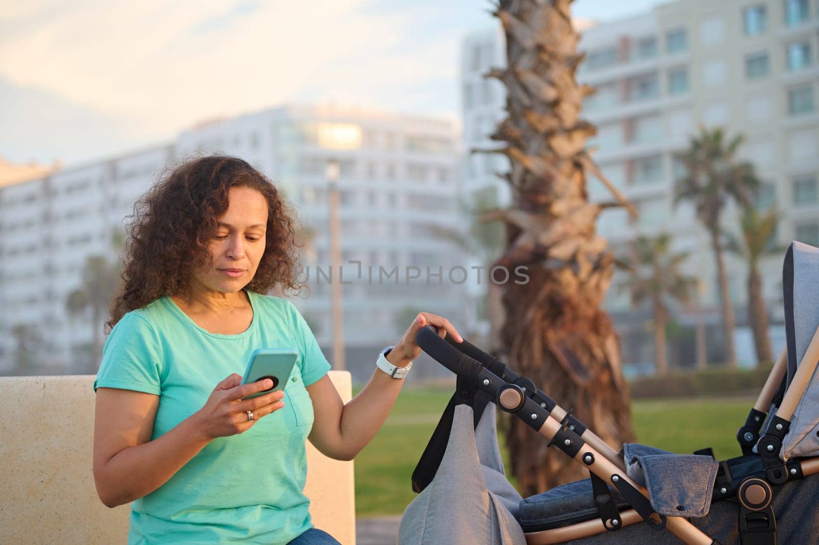 Beautiful young happy woman mother holding using a smart mobile phone, sitting on city bench and pushing baby pram on the promenade at sunset. People. Maternity leave lifestyle. Online communication