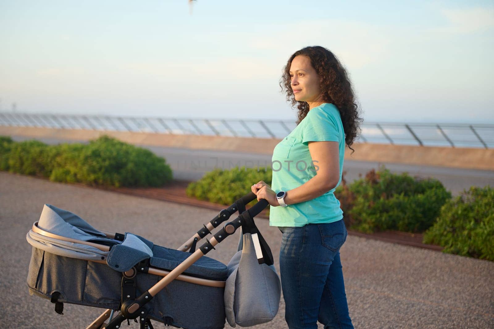 Curly haired multi ethnic brunette woman walking with her baby sleeping in pram, standing on the footpath on the promenade against Atlantic ocean on the background at sunset