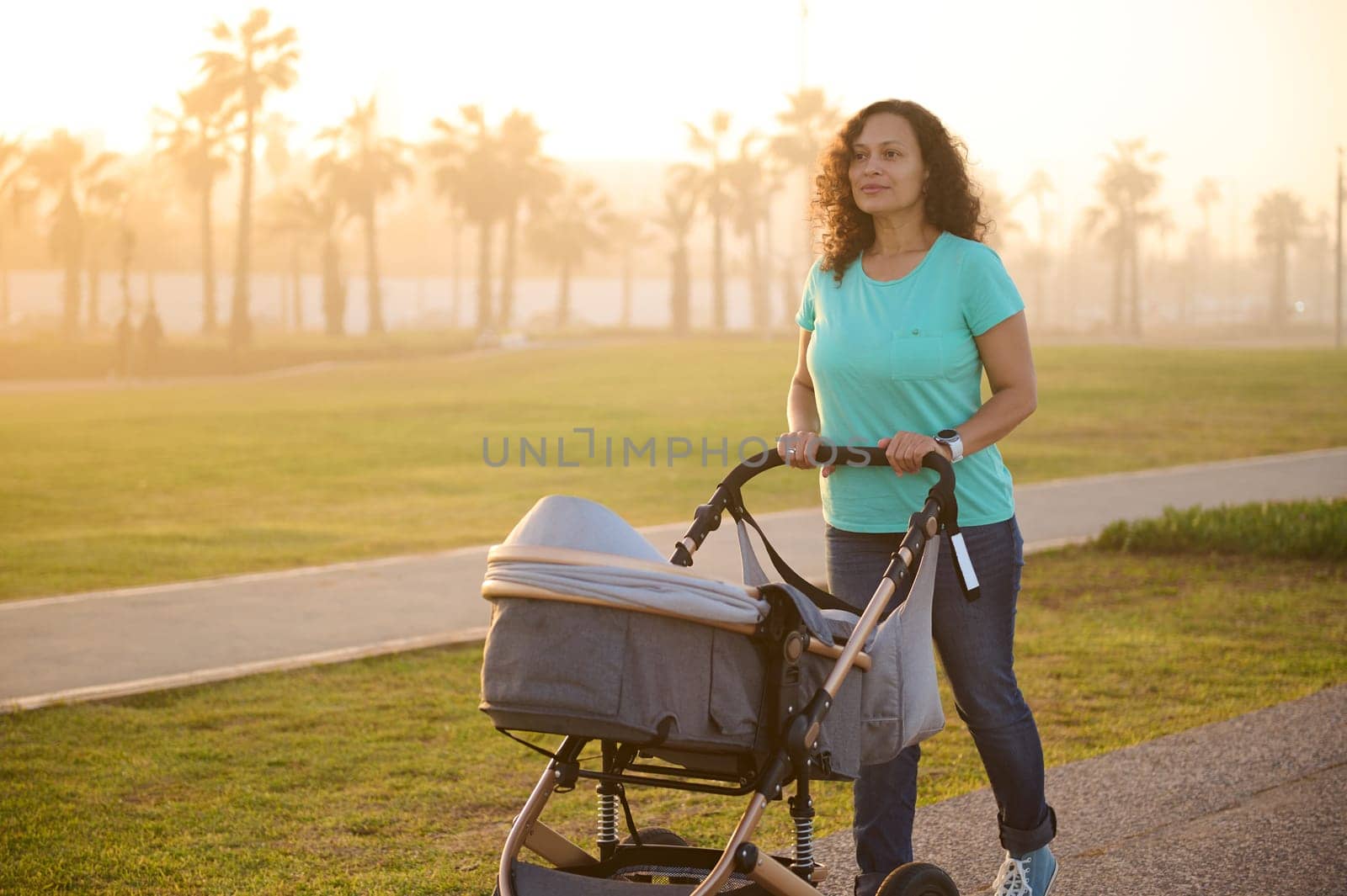 Full size shot of a happy loving mother looking at camera, pushing a baby pram while walking on the Atlantic ocean promenade at sunset. People. Maternity leave lifestyle. Motherhood and infancy.