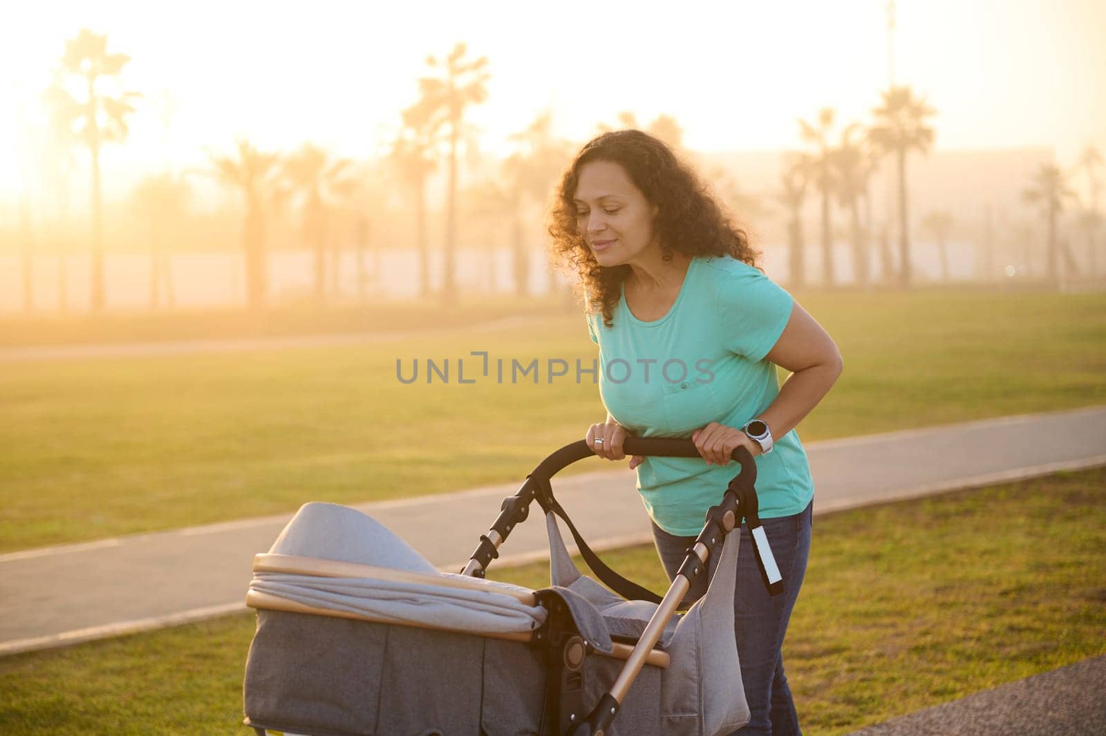 Mum walking on city street at sunset. Multi ethnic cheerful young adult woman pushing her newborn baby sleeping in a pram, during daily outdoors walking. Family concept and healthy lifestyle