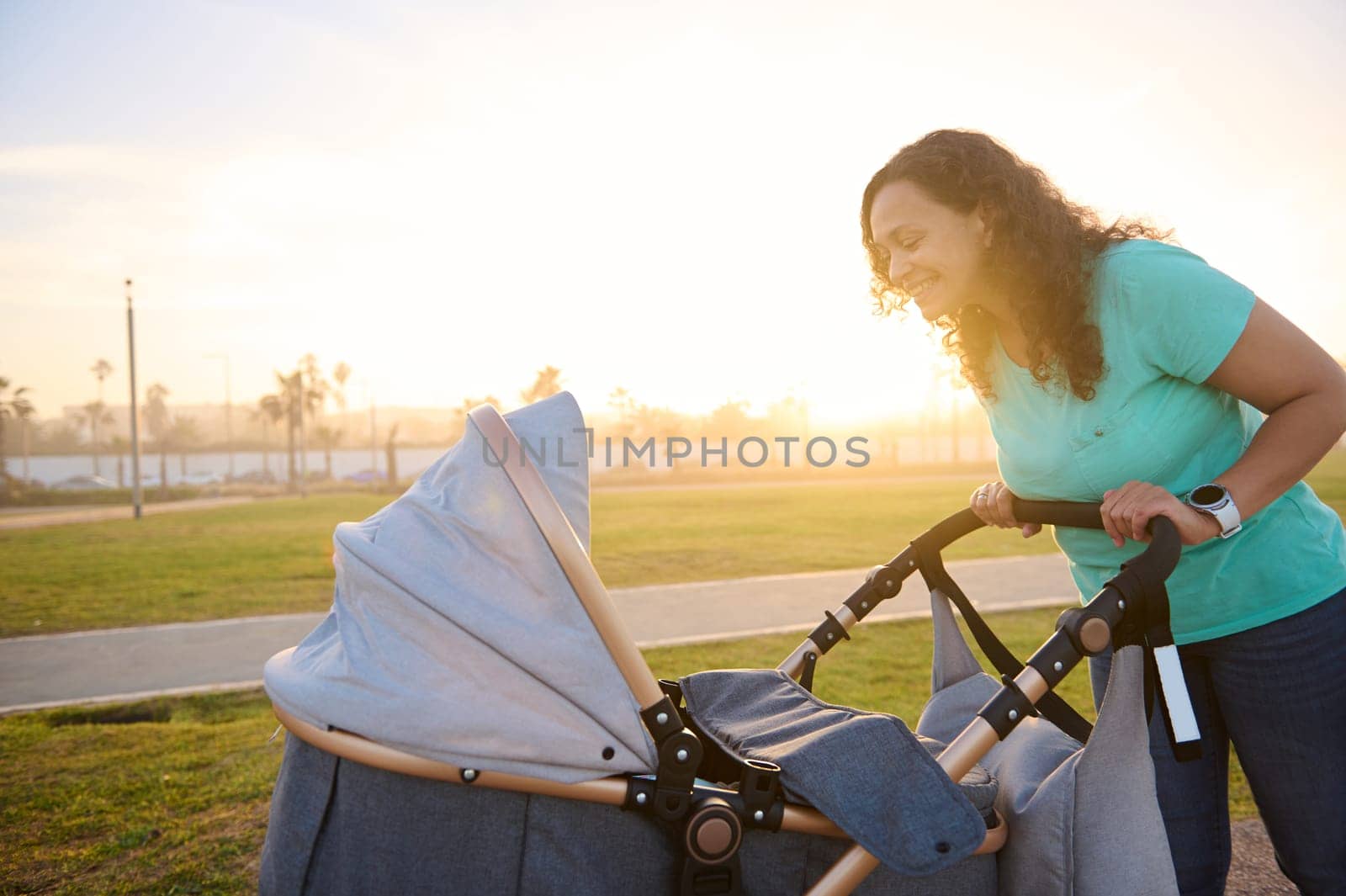 Side portrait of authentic happy mother looking and pushing infant baby stroller while walking in park at sunset. The concept of healthy active lifestyle. Daily outdoors walking and maternity leave.