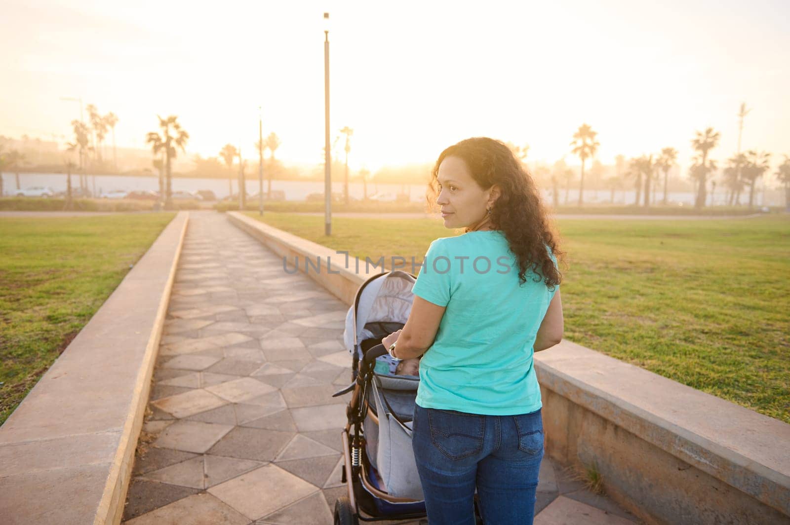 Rear view of a smiling beautiful and young woman 40s, loving caring mother and newborn baby in a stroller walking in the park at sunset background. Copy space