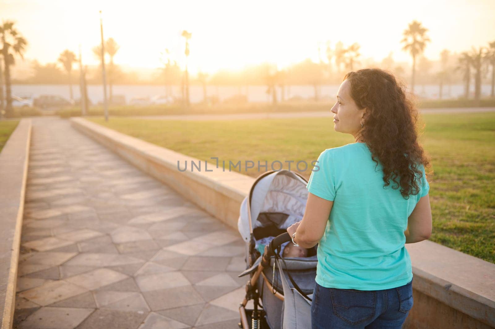 Rear view of happy smiling young woman 40s, loving caring mother carrying her little child, a newborn baby in his baby troll, enjoying walk on the promenade at beautiful sunset. Copy advertising space by artgf