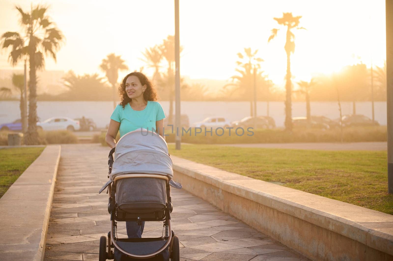 Happy smiling young mother pushing gray silver baby stroller. Daily outside walking, routine. Infant sleeping and breathing fresh air in summer at sunset. Motherhood. Family. Maternity leave lifestyle
