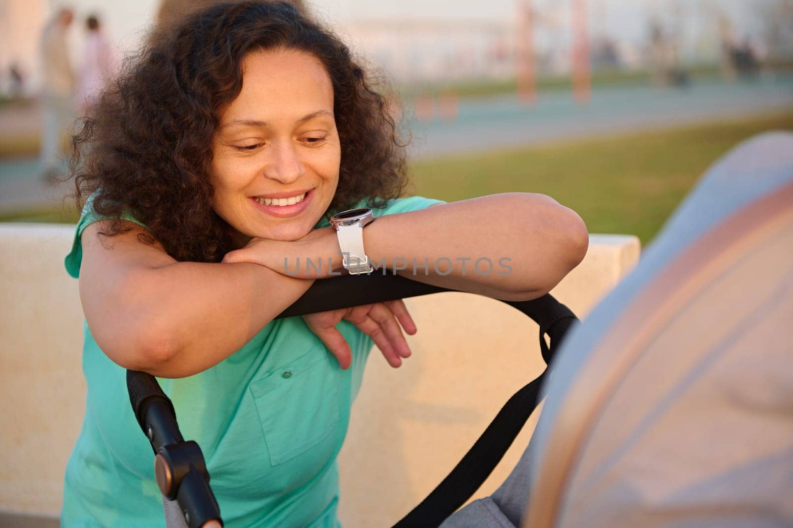 Portrait of happy mother smiling broadly to her baby in the baby pram. Multi ethnic curly haired brunette woman in blue turquoise t-shirt walking with her newborn child sleeping in the baby carriage