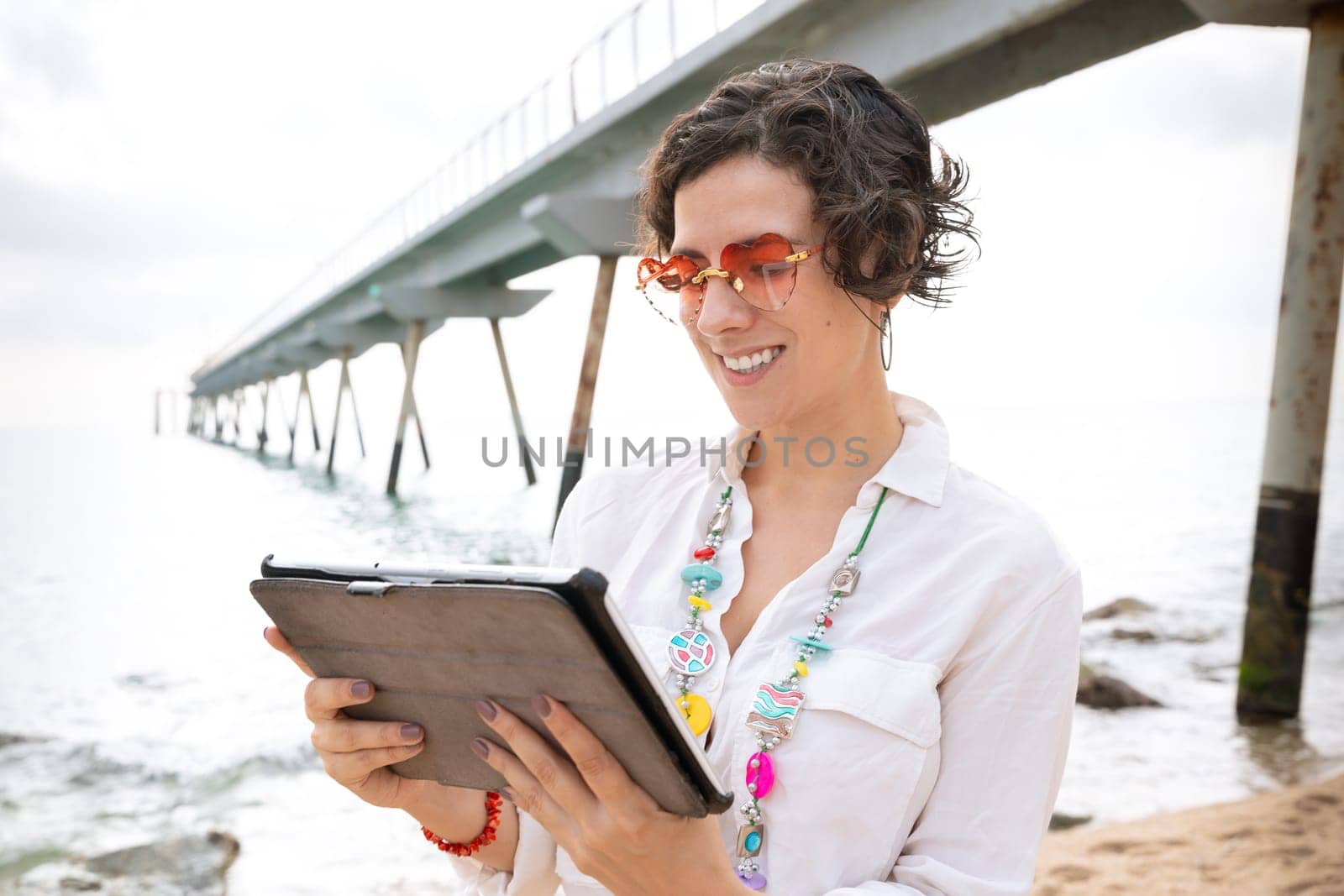 Woman on the beach with digital tablet in hand typing smiling with sunglasses