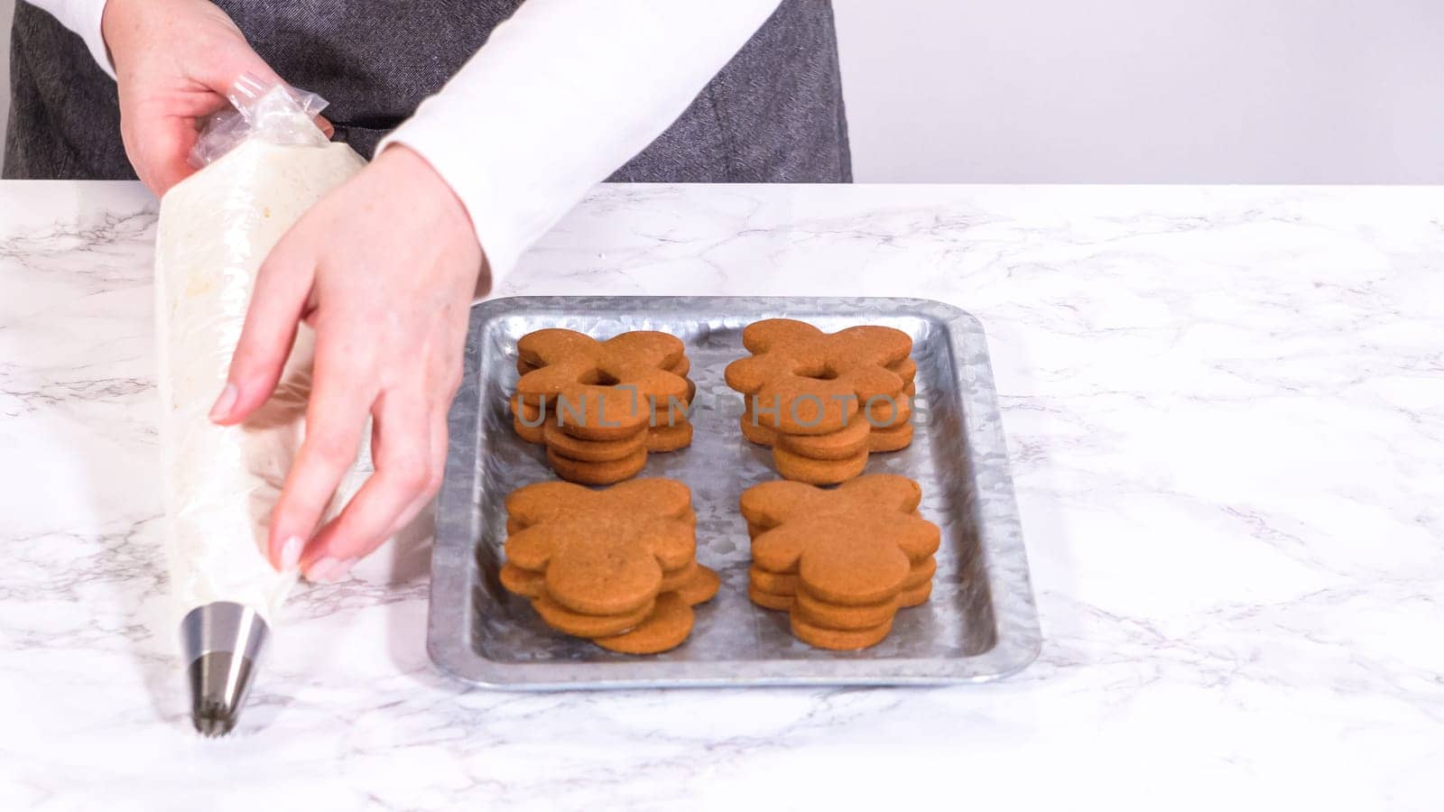 Gingerbread cookies await their second halves on a marble surface, each meticulously piped with buttercream to craft delightful sandwich treats. The precision of the piping adds a festive touch.