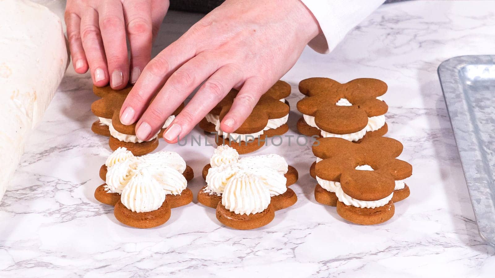 Gingerbread cookies await their second halves on a marble surface, each meticulously piped with buttercream to craft delightful sandwich treats. The precision of the piping adds a festive touch.