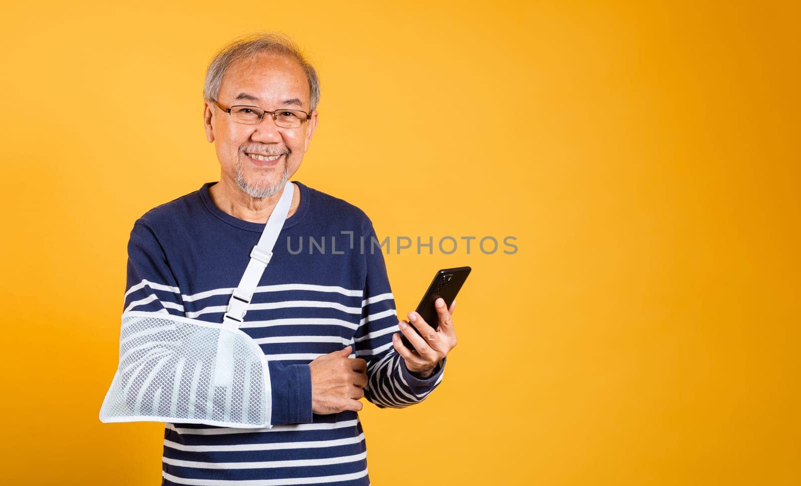 Portrait Asian old man cast his arm and holding mobile phone studio isolated yellow background, retired elderly man accident broken arm wear splint treatment, insurance medical care application