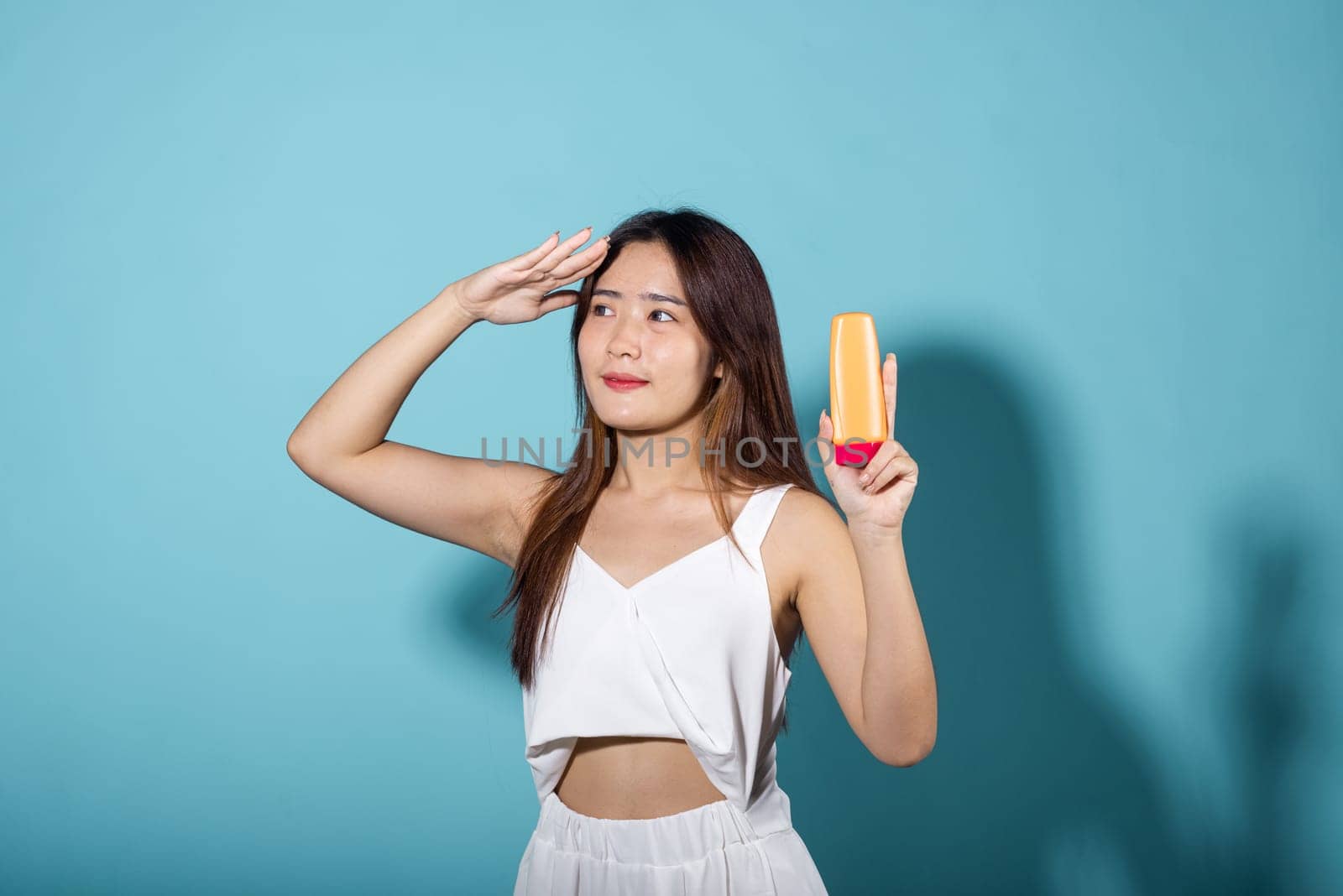 Beautiful Asian young woman holding body lotion on hand in studio shot isolated on blue background, Brunette female hold cream sunblock bottle, tube of sunscreen, Beauty cosmetics facial treatment