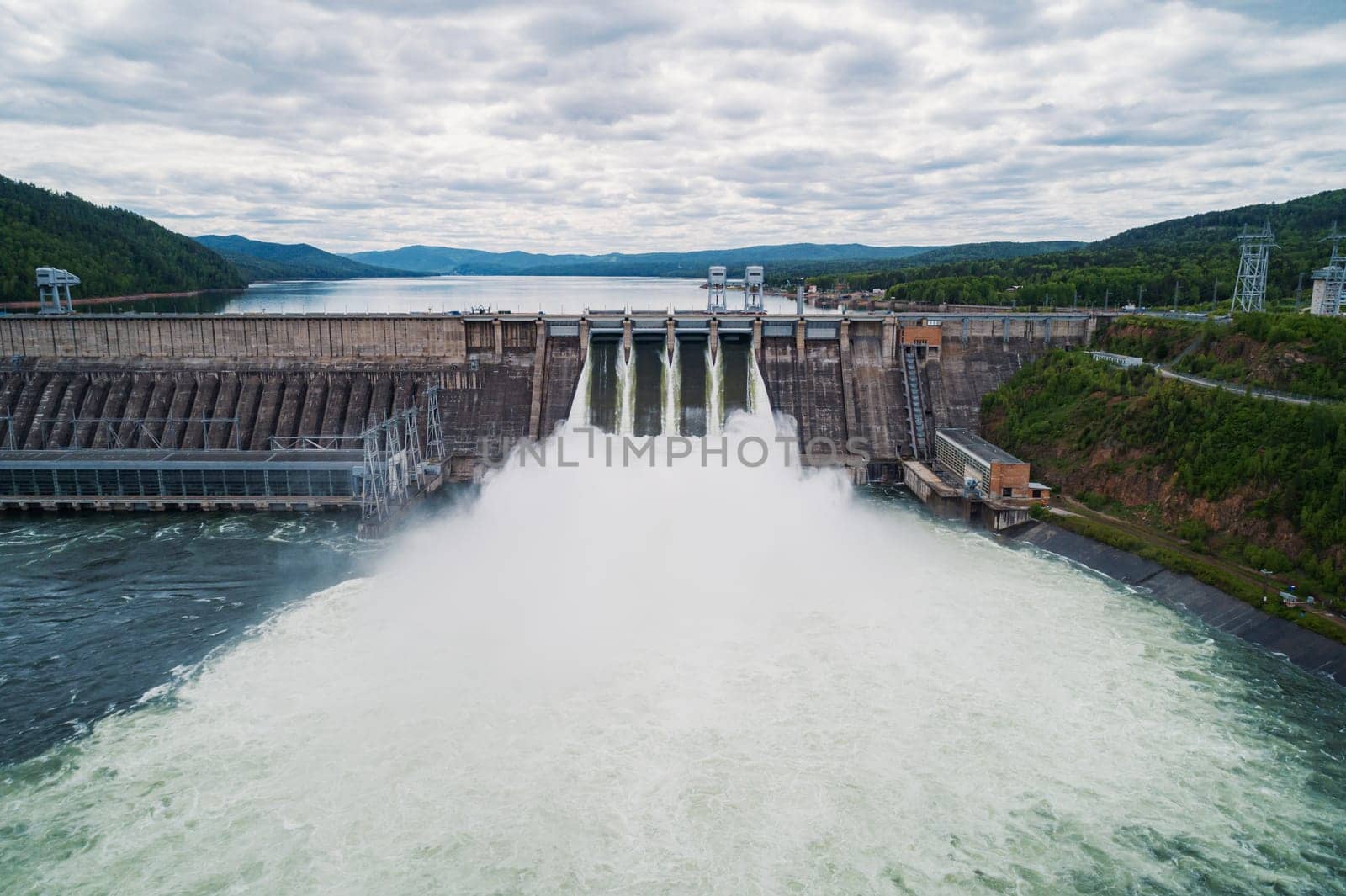 Aerial view of concrete dam releasing water into river on cloudy day. Water discharge at hydroelectric power plant. by Busker