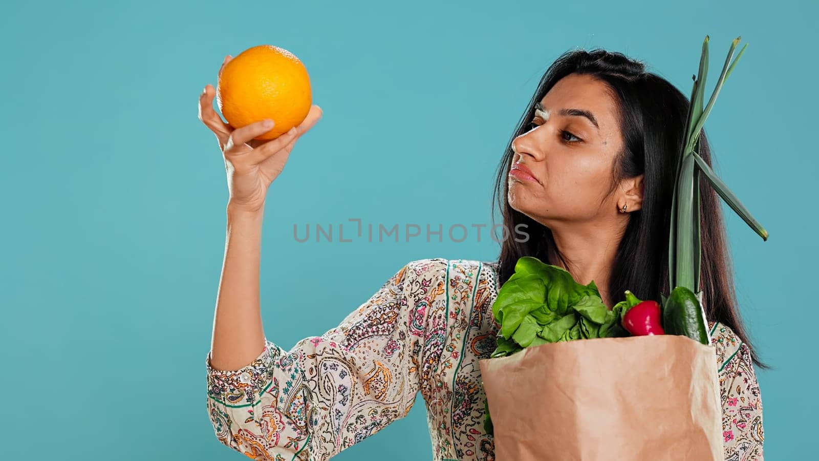 Indian woman with paper bag with vegetables and fruits testing quality, looking at orange, studio background. Vegetarian verifying groceries are ripe after buying them from zero waste shop, camera B