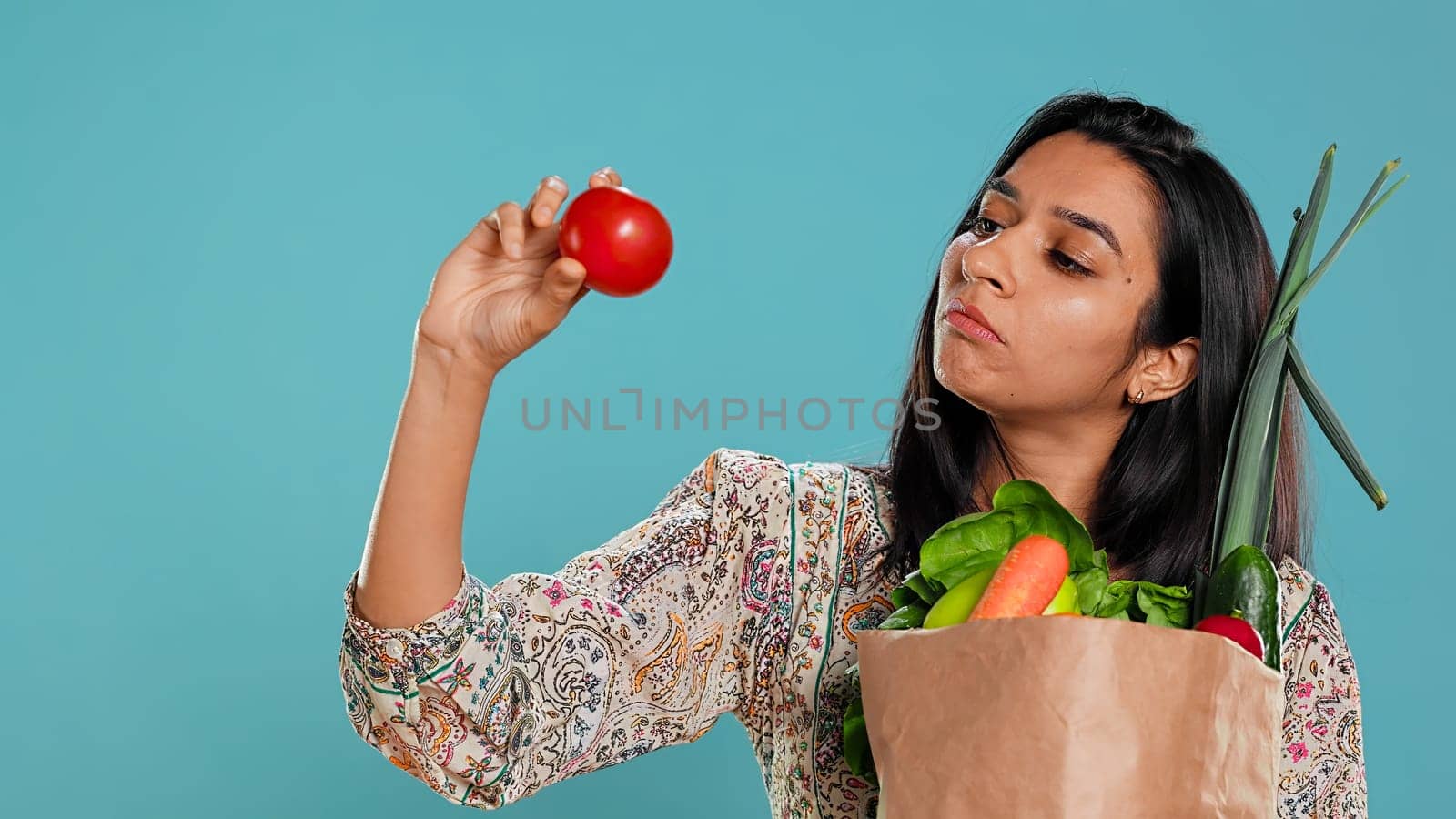 Woman with recycled paper bag in hands filled with vegetables testing quality, studio background. Sustainable living person inspecting groceries after doing purchases in shop, camera B