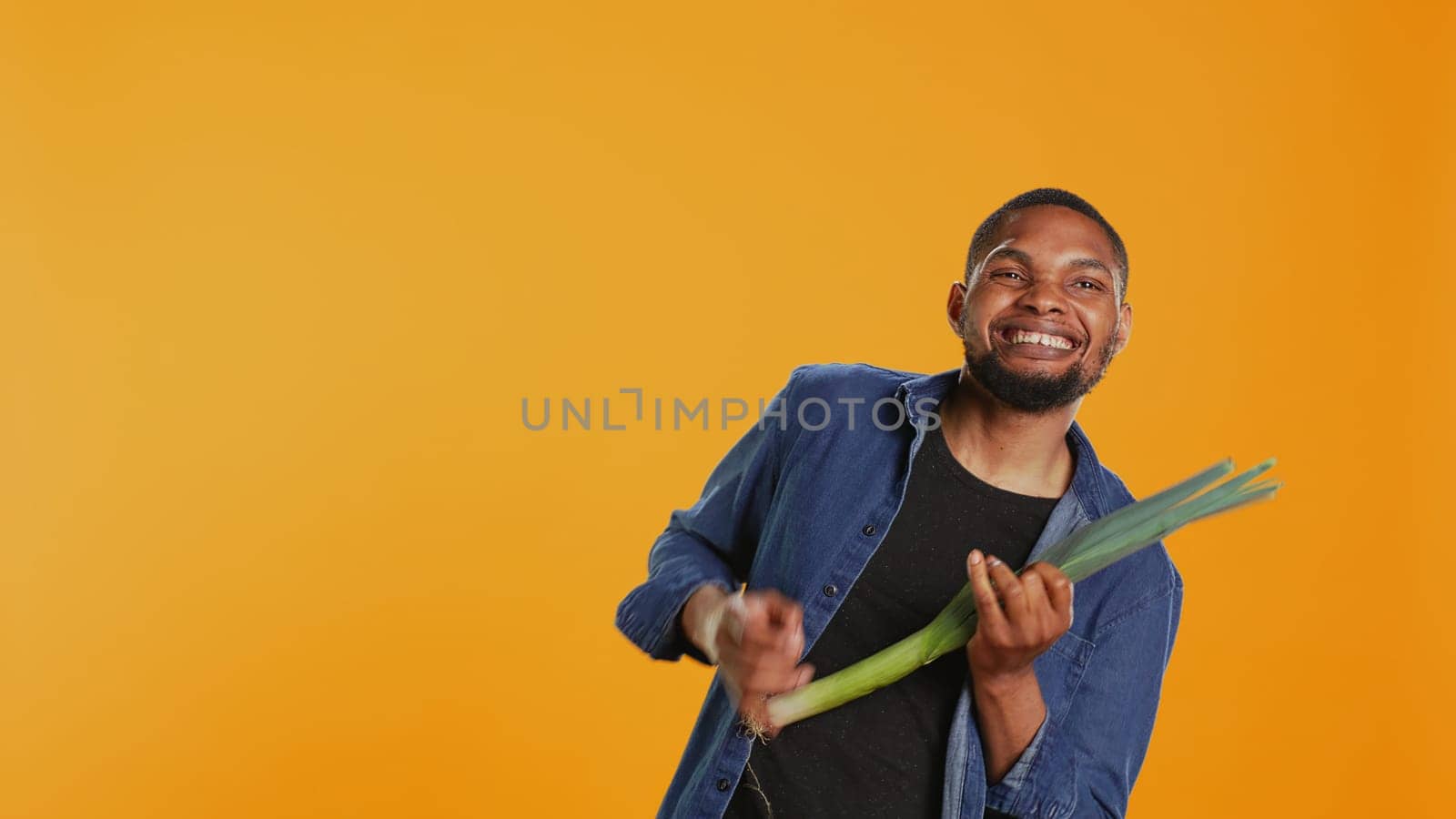 Happy person having fun playing guitar at a leek in studio, acting funny and mimicking a green onion as a musical instrument. Young guy being silly with organic vegetables. Camera B.