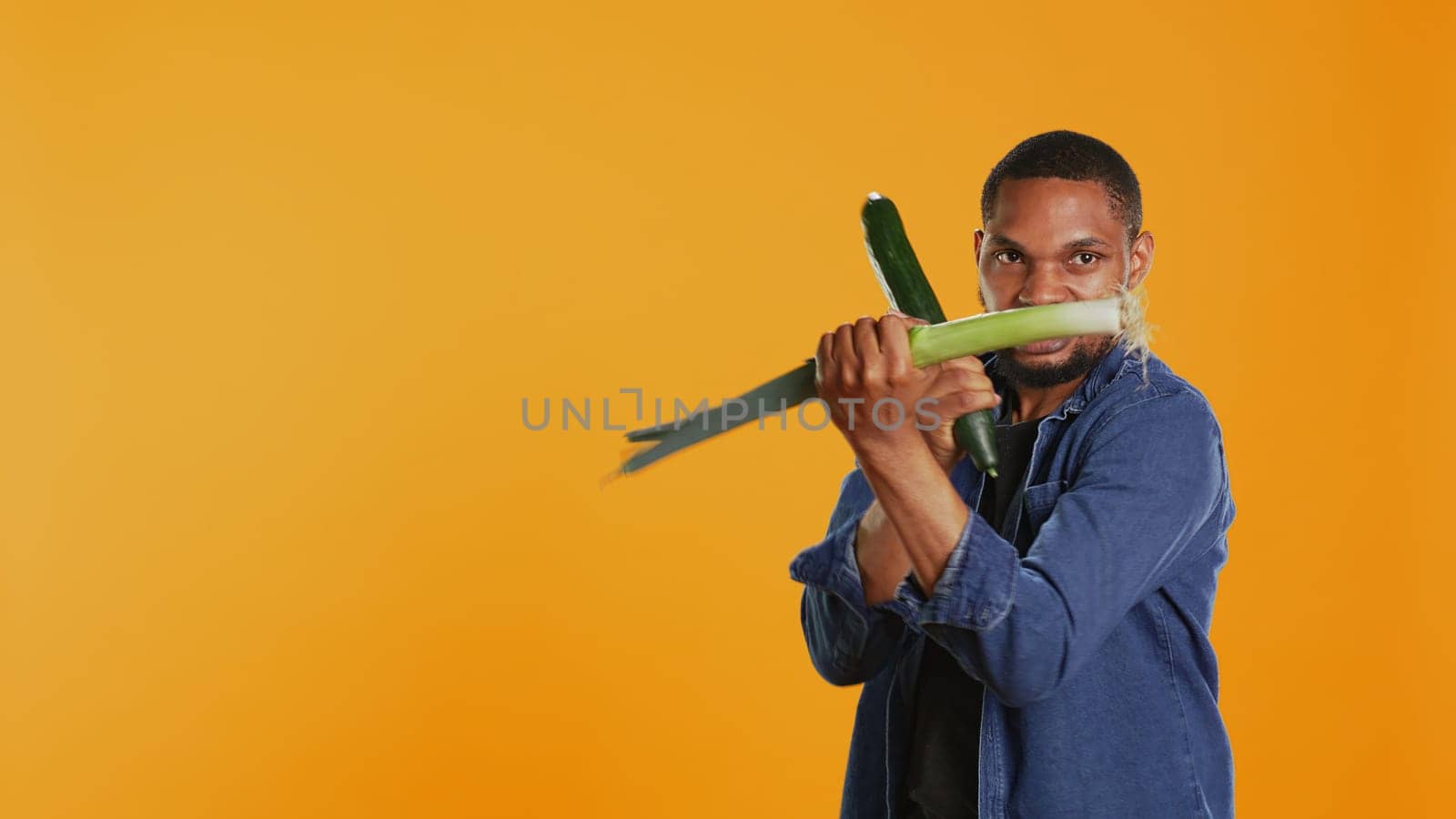 Pleased young adult pretending to start a fight with a cucumber and a leek, acting silly having fun in studio. Vegan guy playing around with natural eco friendly green vegetables. Camera B.