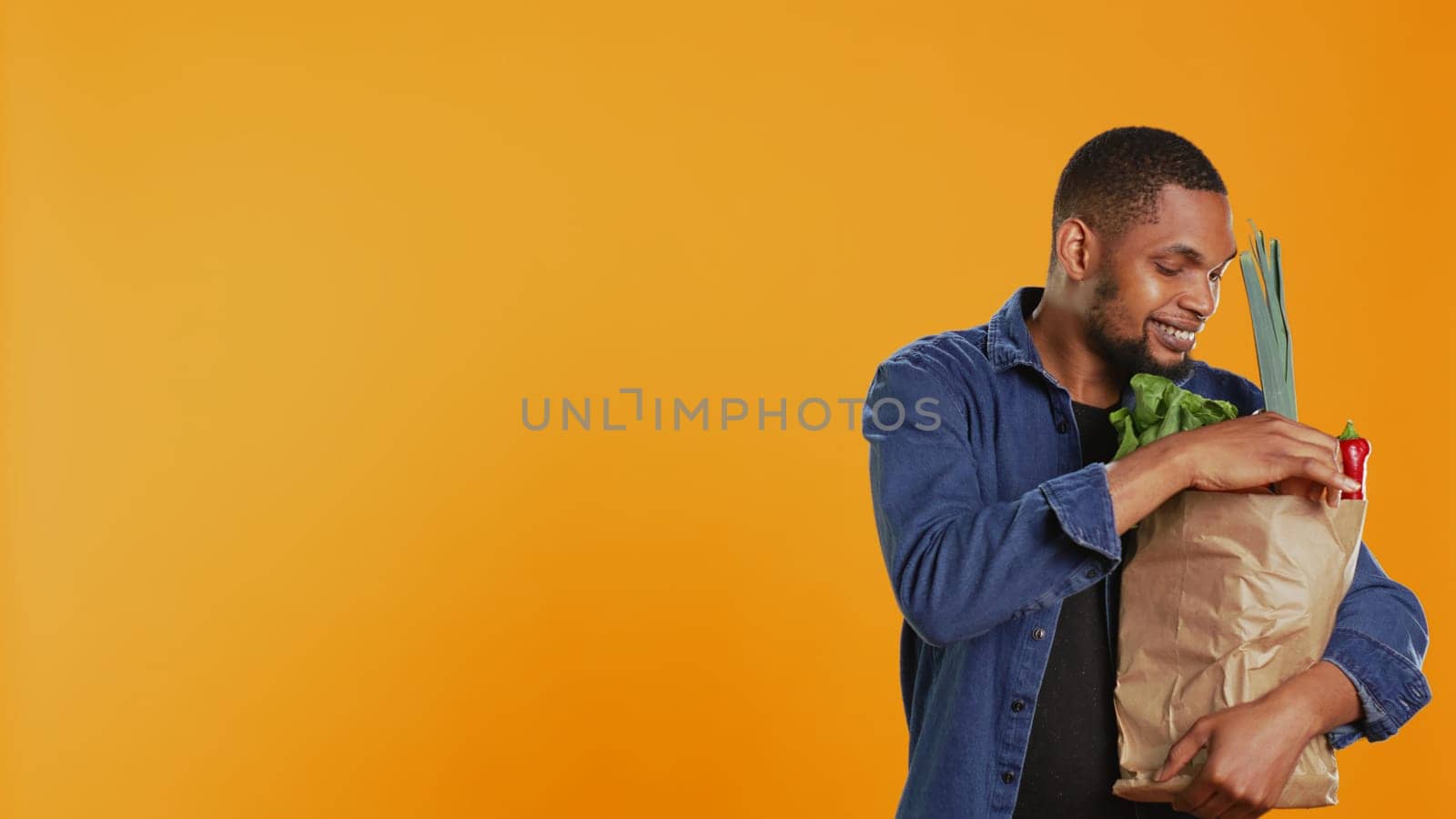 Vegan male person admiring freshly harvested fruits and veggies bought from local farmers market, orange background. Young adult feeling pleased with his bag full of organic goods. Camera B.