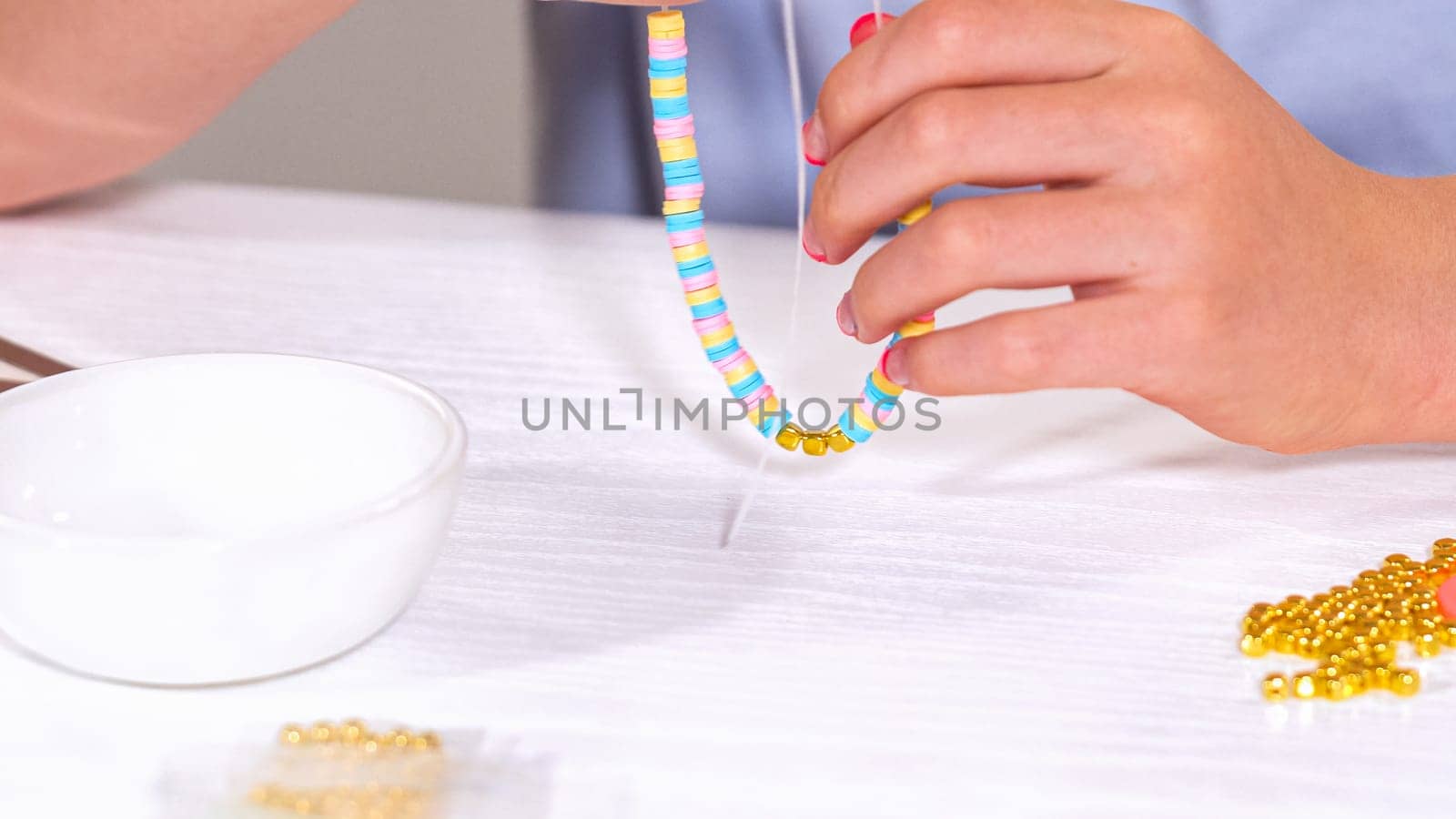 Delicate fingers of a young girl navigate through a treasure trove of bright, multicolored beads, each compartment revealing a new hue to choose from. She's immersed in the joyful task of stringing together a handmade bracelet, with golden beads and pearls lying nearby to add a touch of sparkle to her creation.