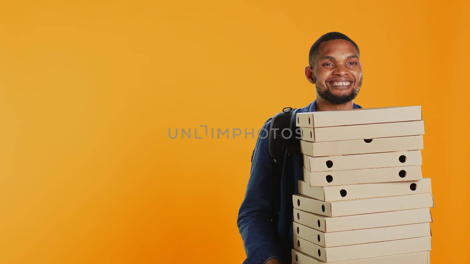 Male pizzeria courier carrying huge pile of pizza boxes in studio, preparing to deliver meal order to customers. Young deliveryman holding a big stack of fast food takeaway, shipping. Camera B.