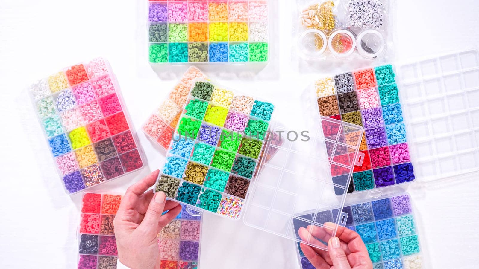 Flat lay. Woman’s hands gracefully poised over a collection of beads, sorted by color in transparent organizers. The array of beads spans a vibrant spectrum, from deep purples to bright oranges, meticulously arranged for easy selection as she embarks on creating a custom piece of jewelry.