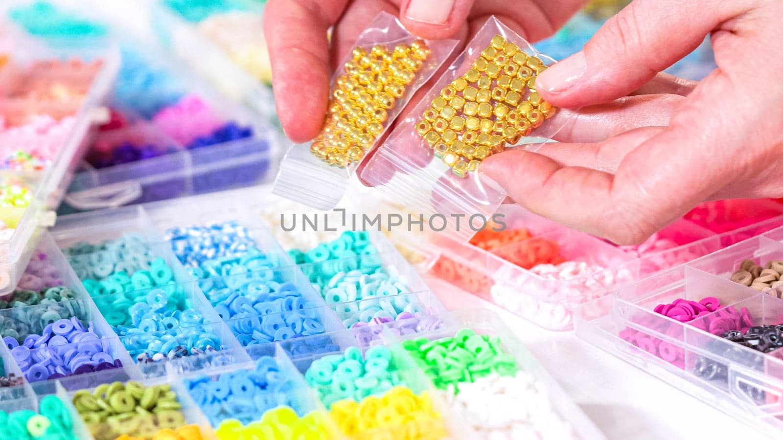 Woman’s hands gracefully poised over a collection of beads, sorted by color in transparent organizers. The array of beads spans a vibrant spectrum, from deep purples to bright oranges, meticulously arranged for easy selection as she embarks on creating a custom piece of jewelry.