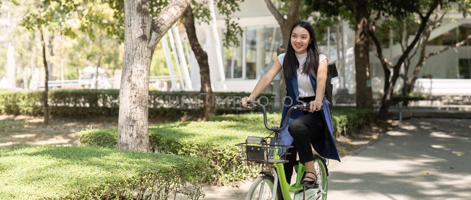 Smiling Asian woman rides a bicycle through a park on a sunny day, promoting eco-friendly transportation