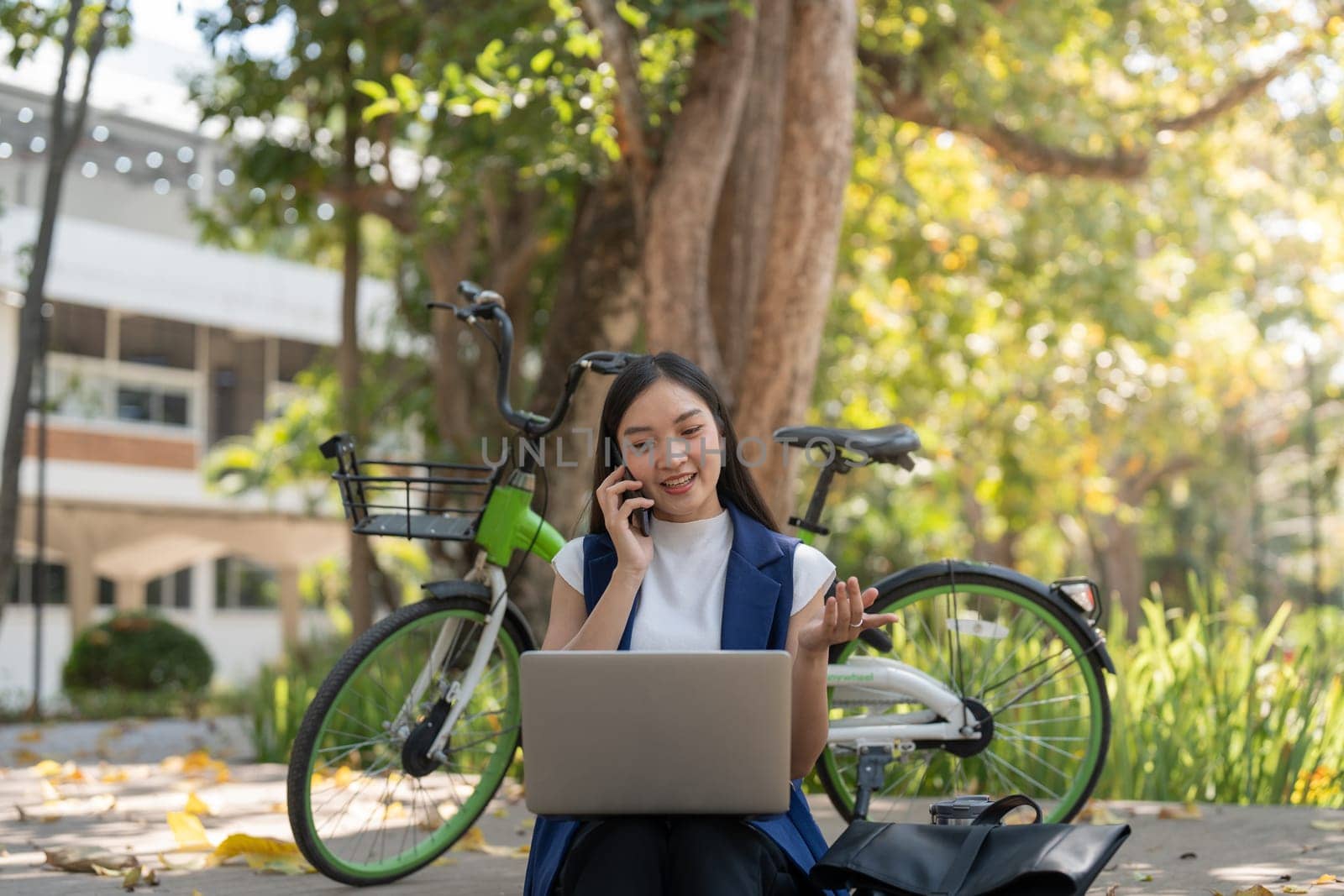 Asian woman sitting outdoors with a laptop and phone, promoting remote work and eco-friendly commuting with a bicycle