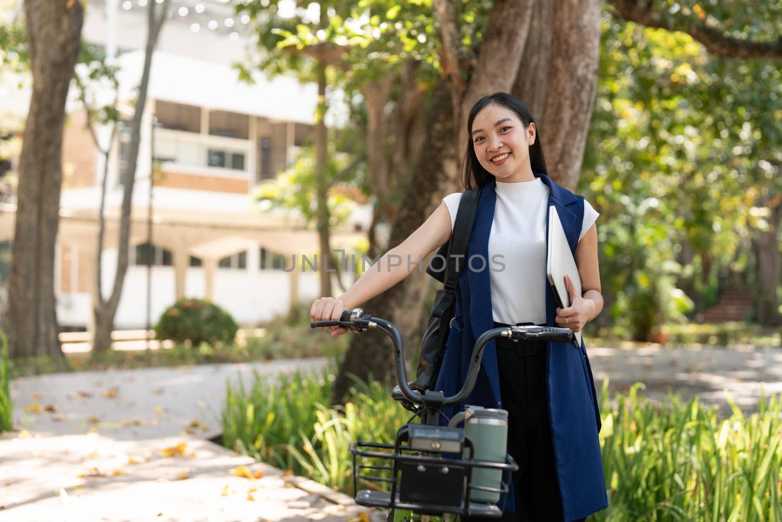Smiling Asian businesswoman holding a laptop and standing with her bicycle outdoors, promoting eco-friendly commuting and professional success