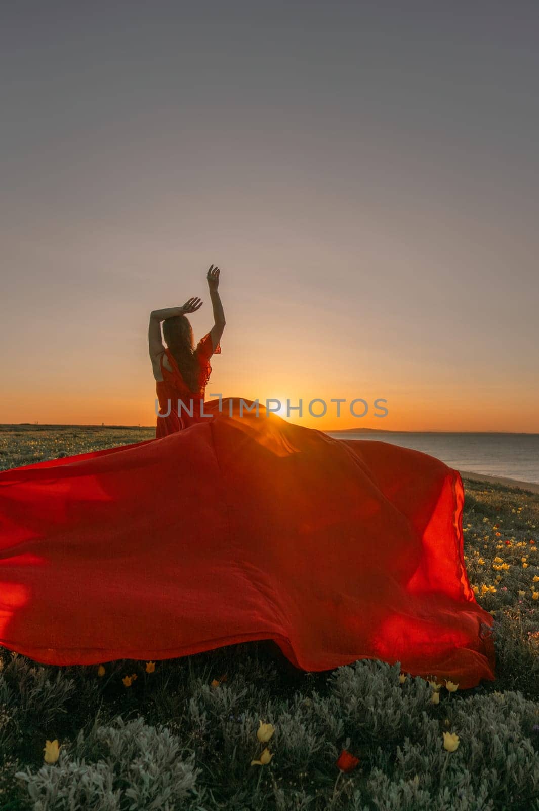 A woman in a red dress is standing in a field with the sun setting behind her. She is reaching up with her arms outstretched, as if she is trying to catch the sun. The scene is serene and peaceful