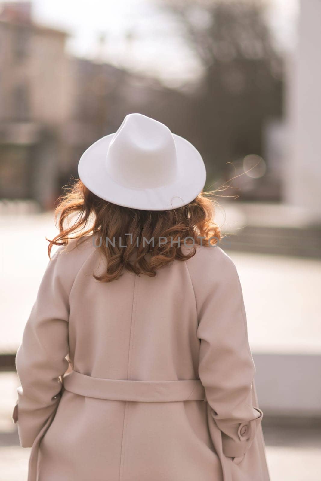 Back view of happy woman wearing hat and coat walking down street on sunny spring day. People, lifestyle, travel and vacations concept by Matiunina