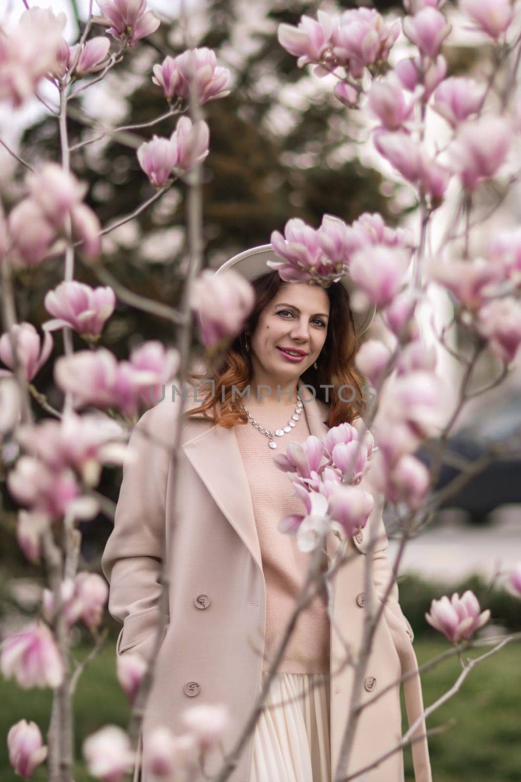 Woman magnolia flowers, surrounded by blossoming trees, hair down, white hat, wearing a light coat. Captured during spring, showcasing natural beauty and seasonal change