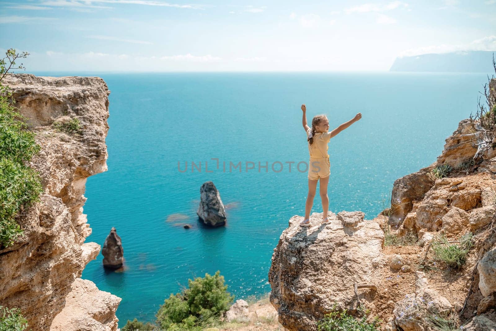 Happy girl stands on a rock high above the sea, wearing a yellow jumpsuit and sporting braided hair, depicting the idea of a summer vacation by the sea