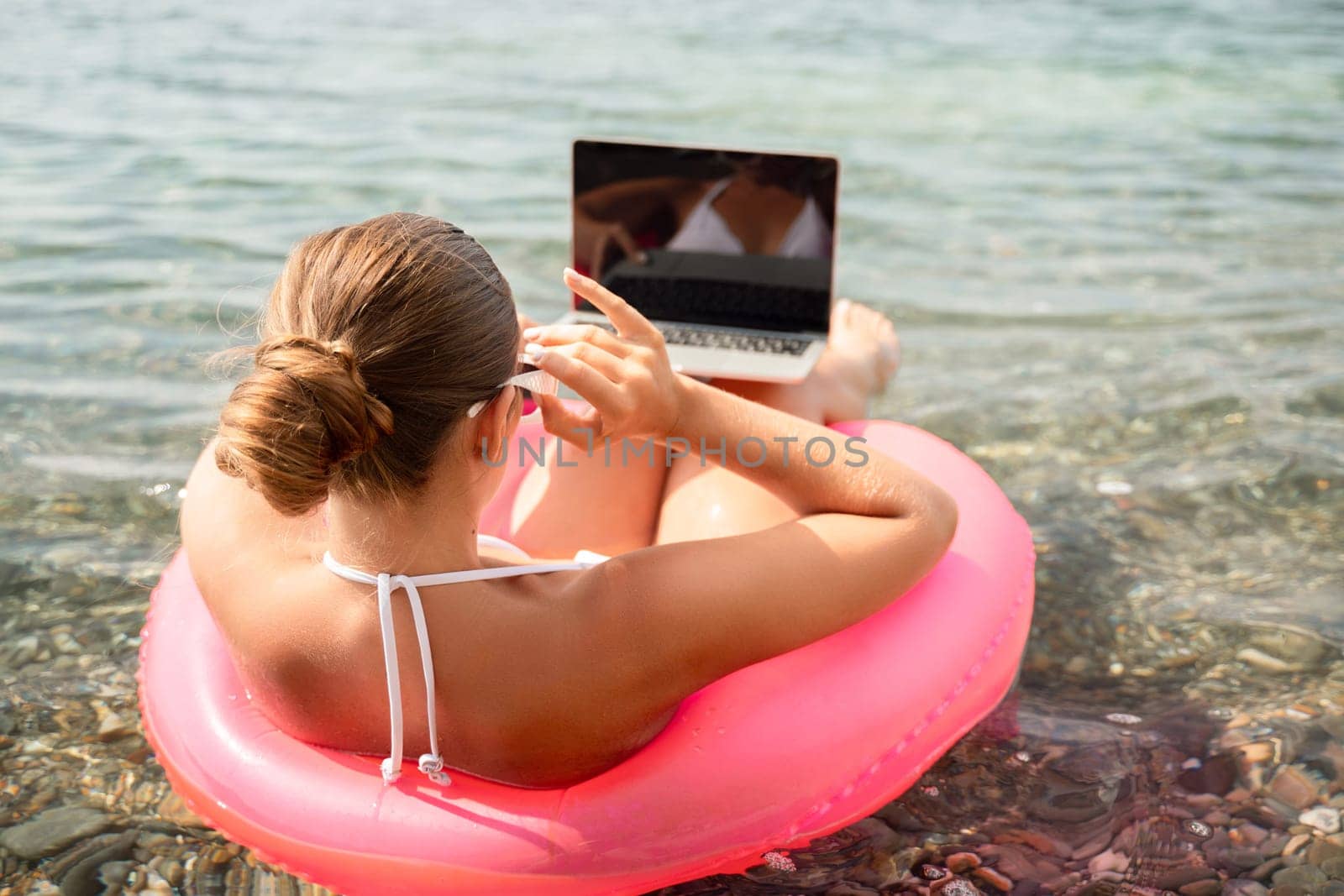 A woman is sitting on a pink inflatable raft in the water, using a laptop. Concept of relaxation and leisure, as the woman is enjoying her time by the water while working on her laptop