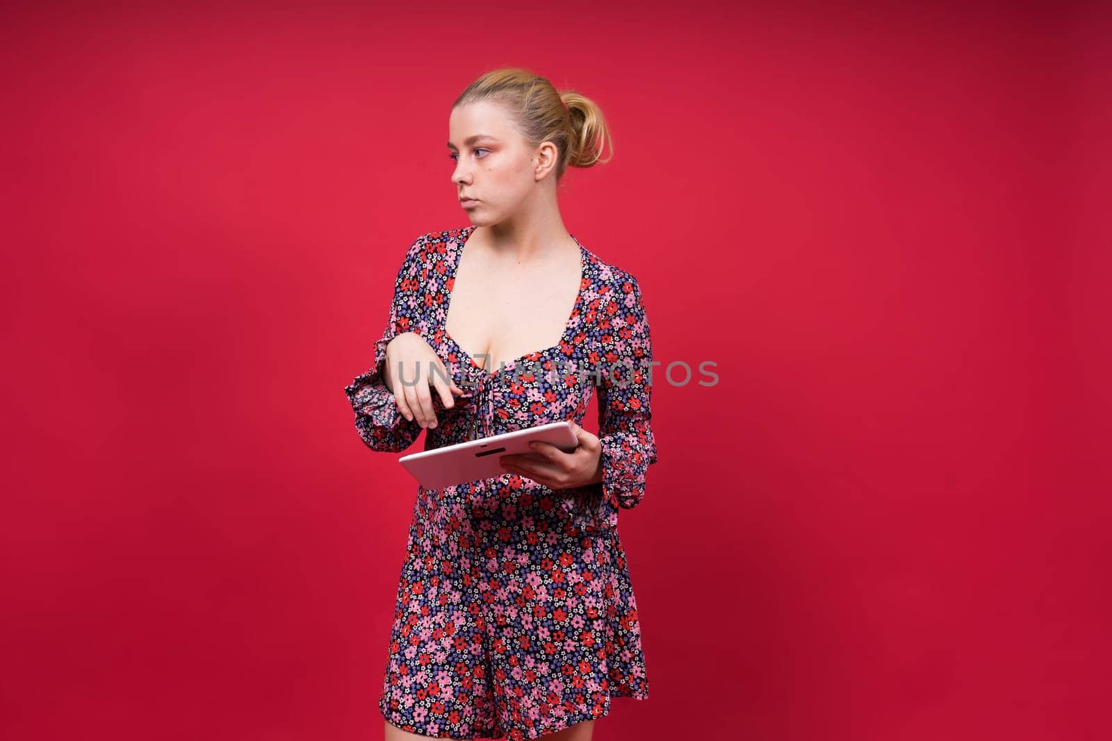 A woman in a purple floral dress is holding a tablet computer, her arm raised in a gesture. She has a microphone pinned to her onepiece garment, with lipstick and flash photography visible