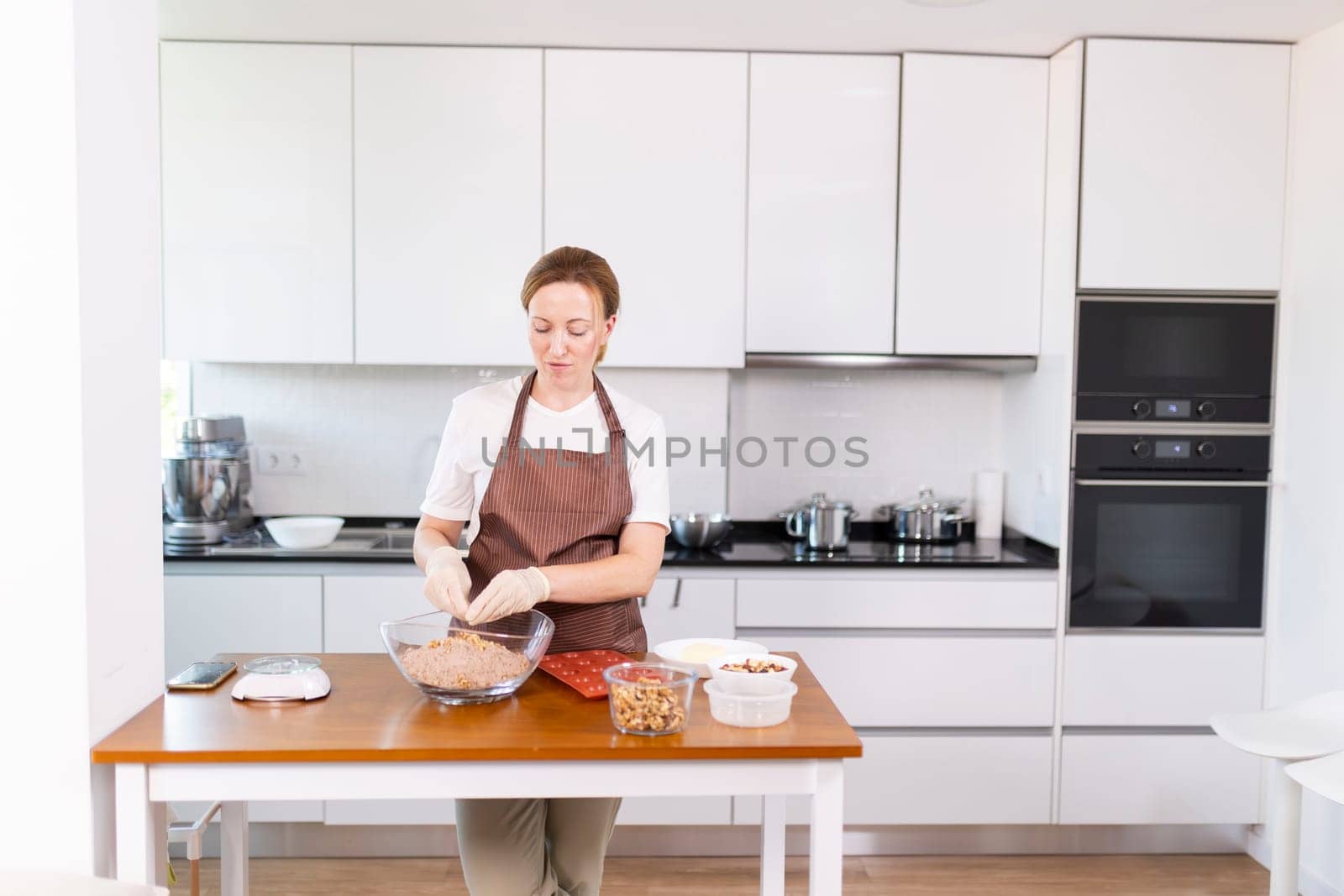 A woman is standing in a kitchen, preparing food. She is wearing an apron and has a bowl of food in front of her. The kitchen is clean and well-organized, with various appliances such as an oven