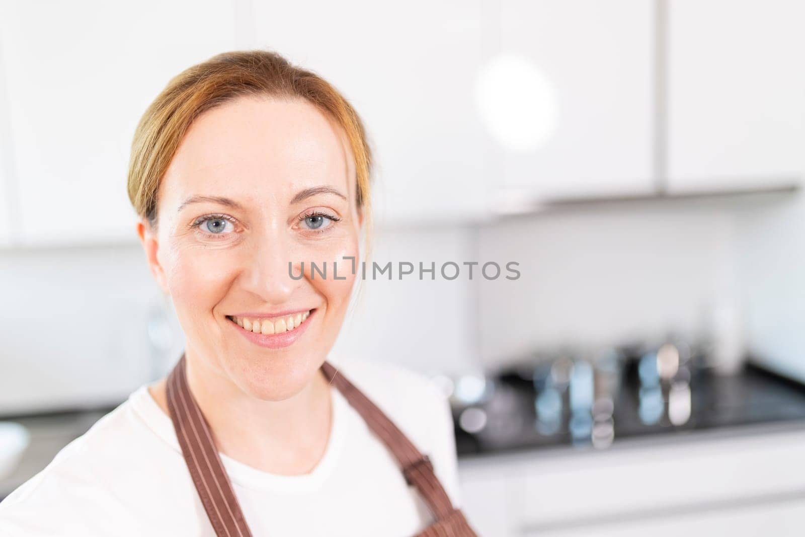 A woman with blonde hair and blue eyes is smiling in front of a kitchen counter. She is wearing an apron and a white shirt