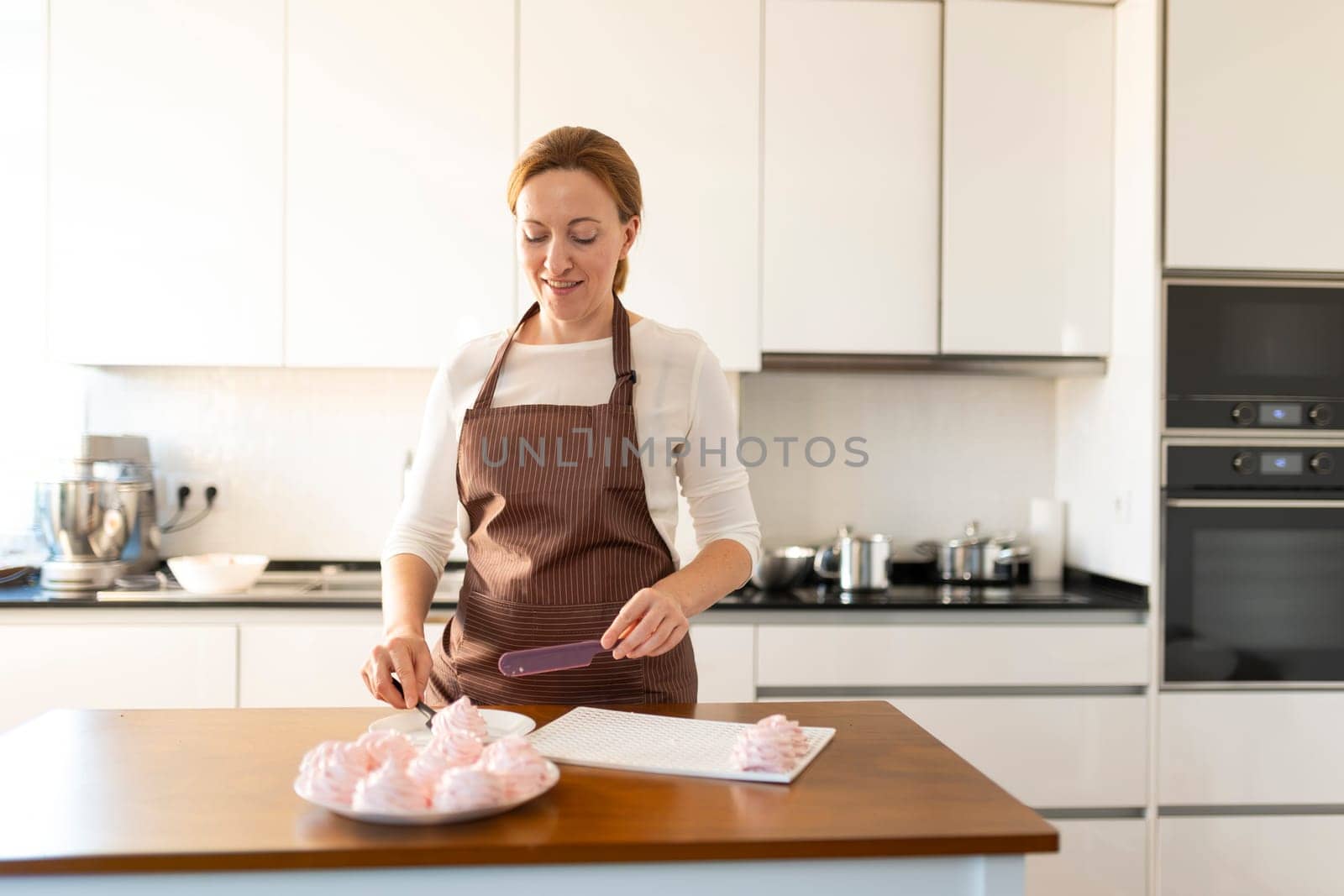 A woman is cutting a dessert on a wooden table. She is wearing an apron and a brown shirt. The dessert is a pastry, and there are several of them on the table