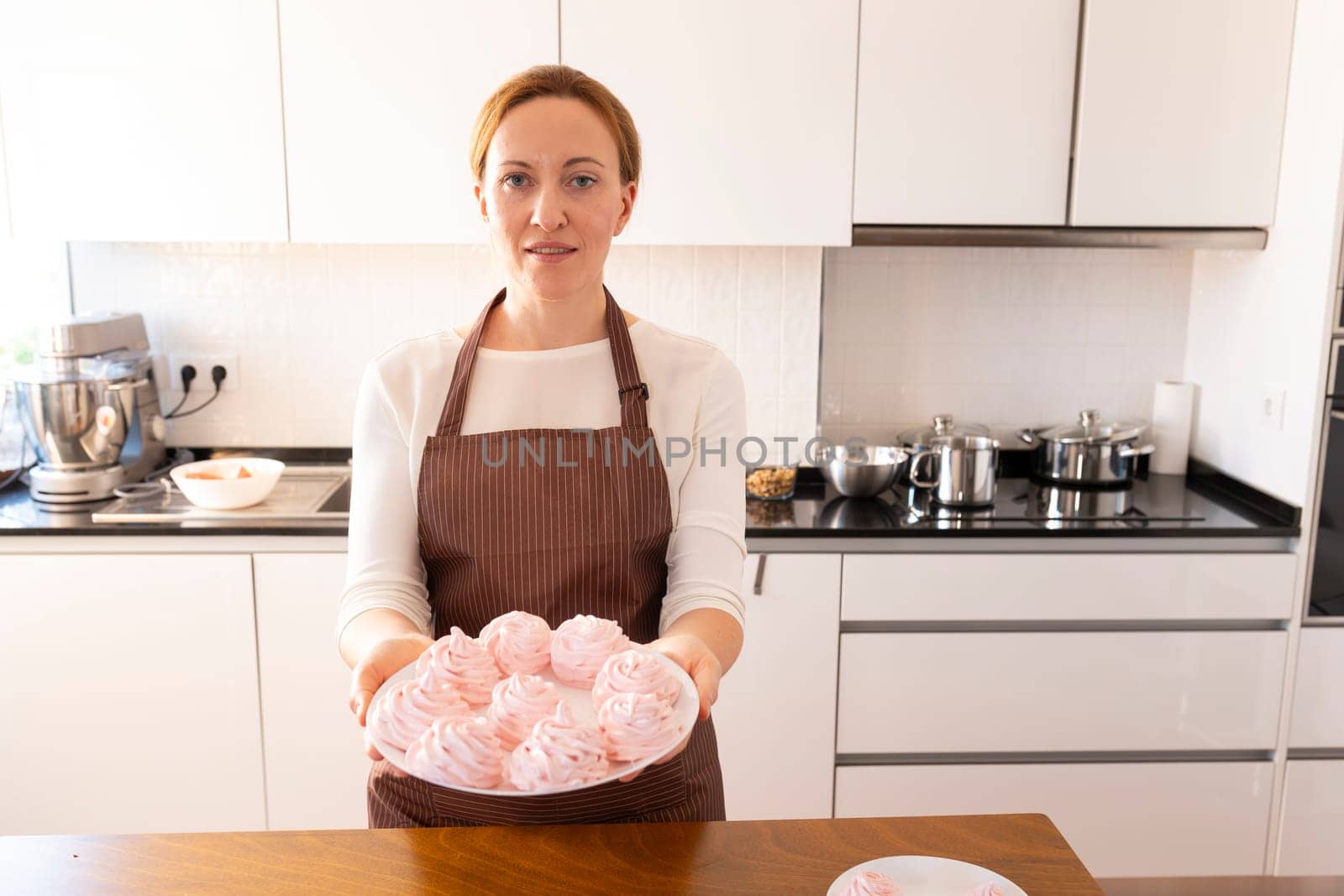 A woman is holding a plate of cupcakes in a kitchen. She is wearing an apron and smiling