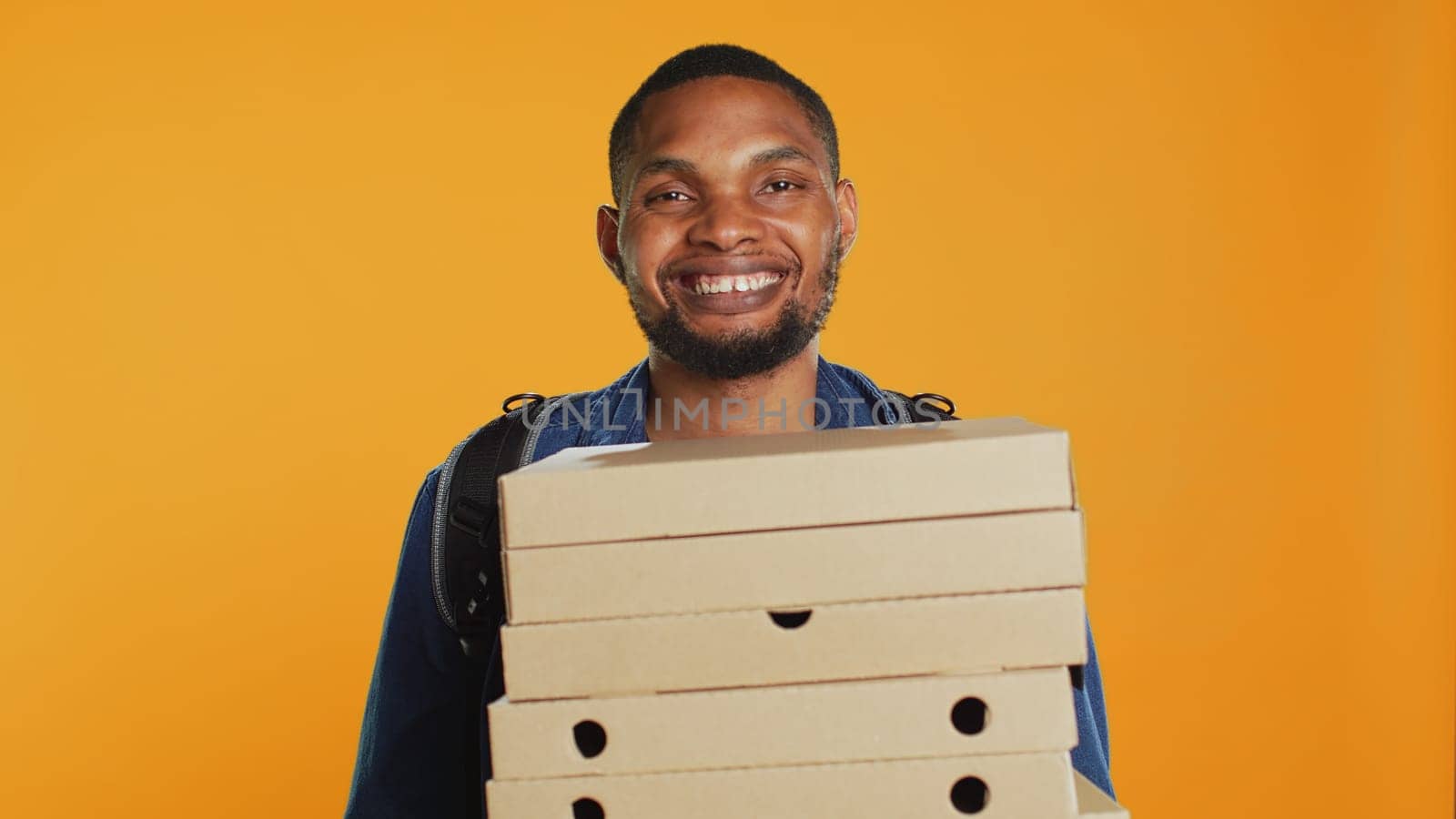 African american pizzeria deliveryman holding stack of pizza boxes to deliver food order to customer in studio. Friendly takeout service employee carries takeaway meal package. Camera A.