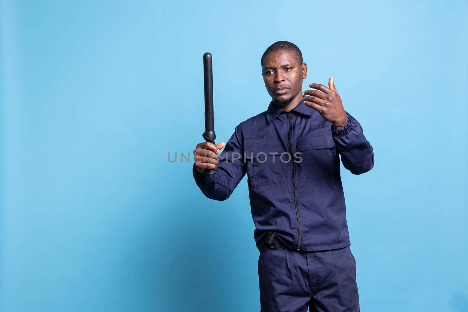 Security agent holding a baton and asking people to come over, calling persons to approach him against blue background. Skilled trained bodyguard working on public safety, job duty.