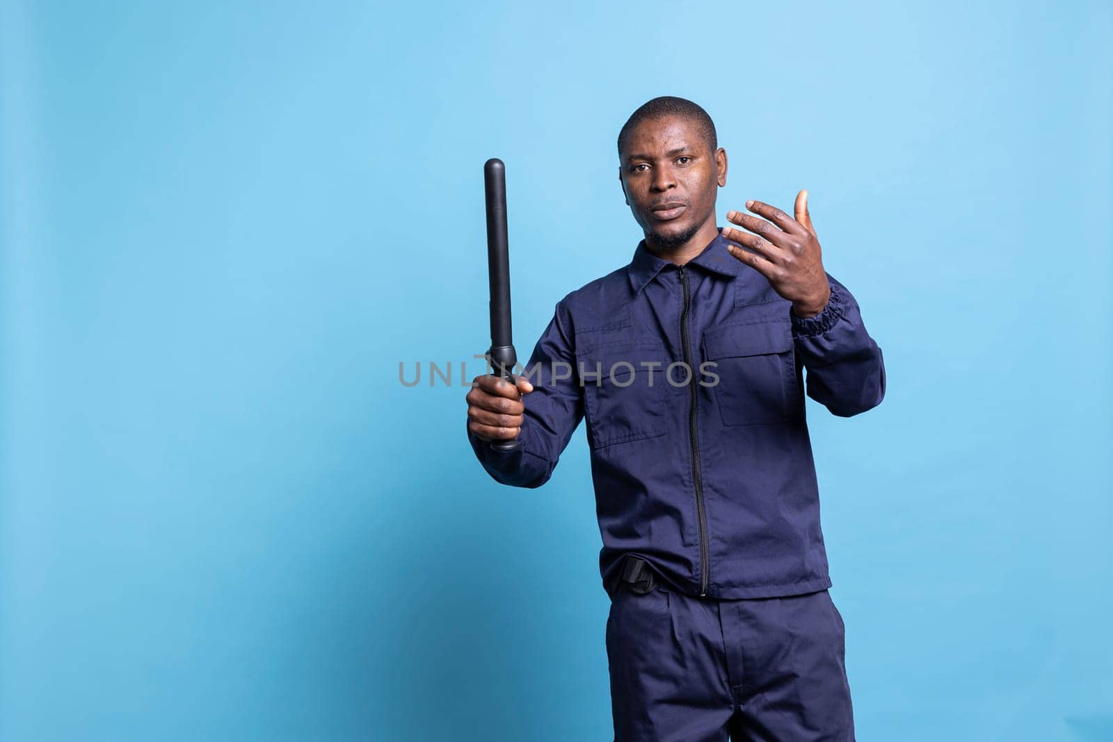 Security guard with a baton in his hand calling people over, asking persons to approach him against blue background. Young armed patrol agent working on public protection and safety.