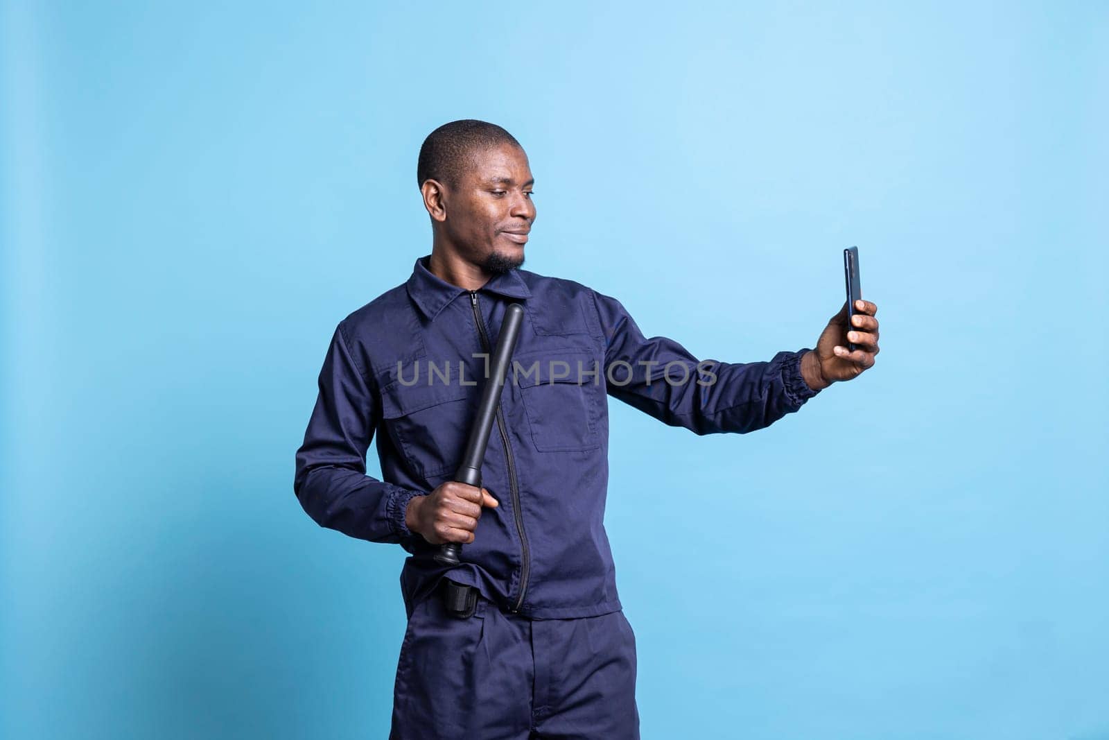 Smiling security agent taking photos with his baton on camera, using a smartphone to take pictures against blue background. Young guard makes memories in his patrol uniform in studio.