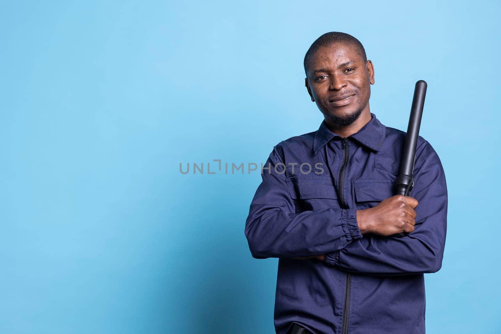 African american security officer feels proud with his duty in studio, posing with confidence holding his baton dressed in professional uniform. Skilled bodyguard standing against blue background.