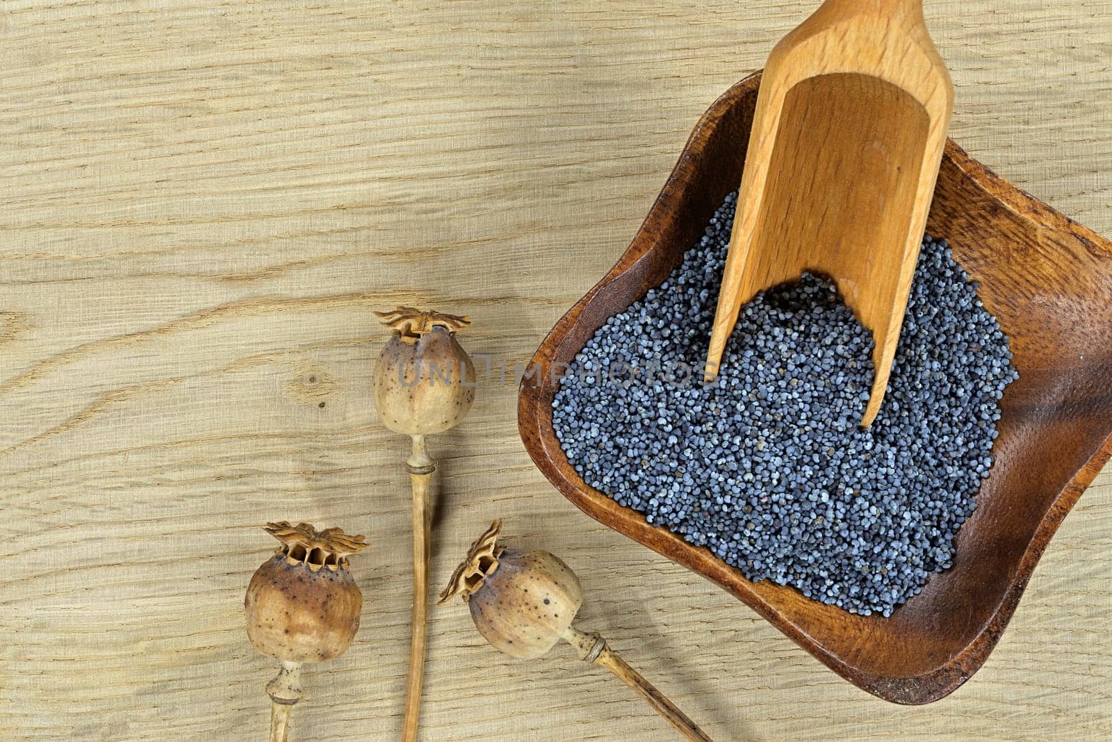 Small wooden bowl filled with black poppy seeds and dried poppy seed heads on rustic wooden table