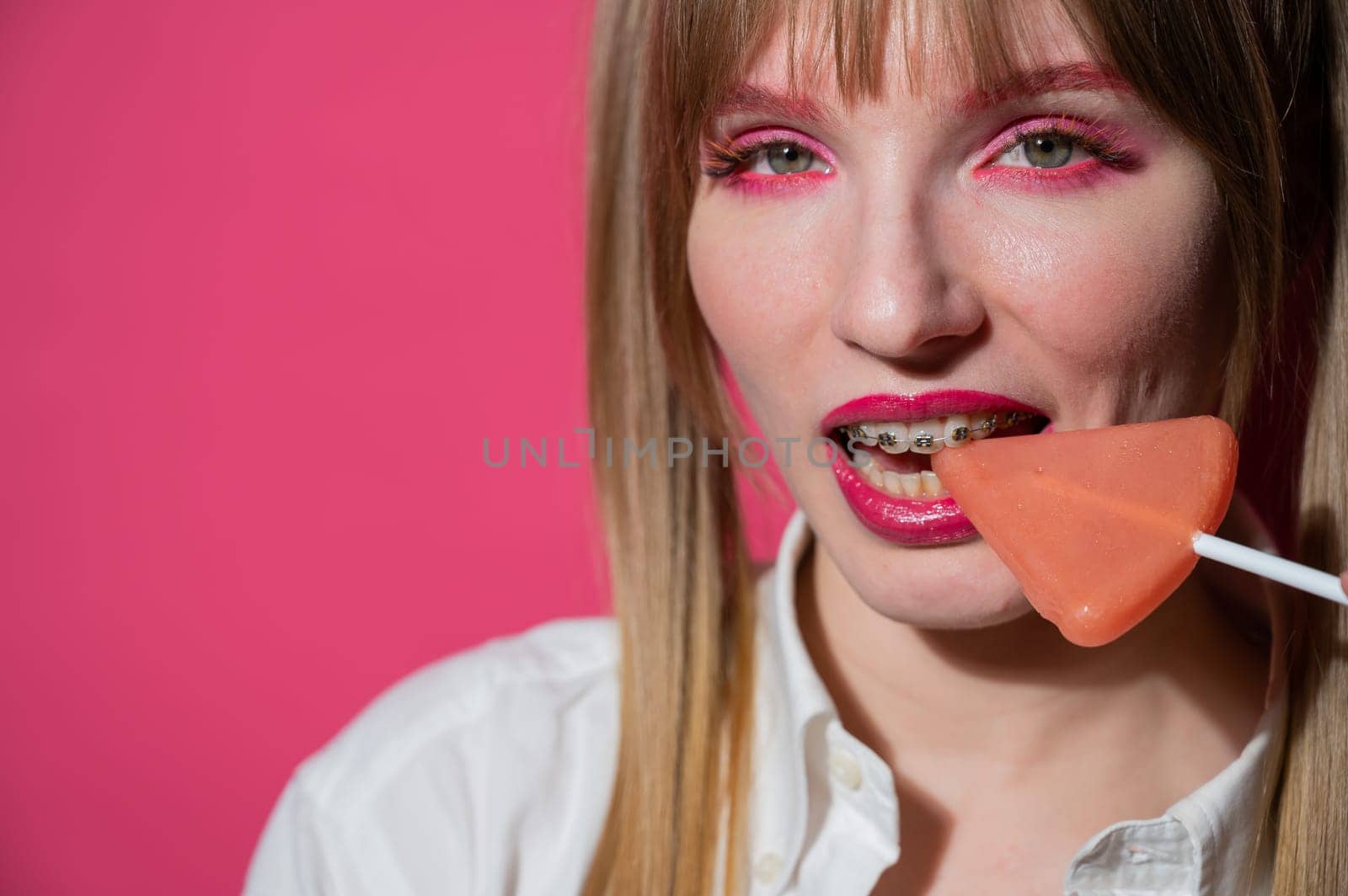 Portrait of a young woman with braces and bright makeup eating a lollipop on a pink background