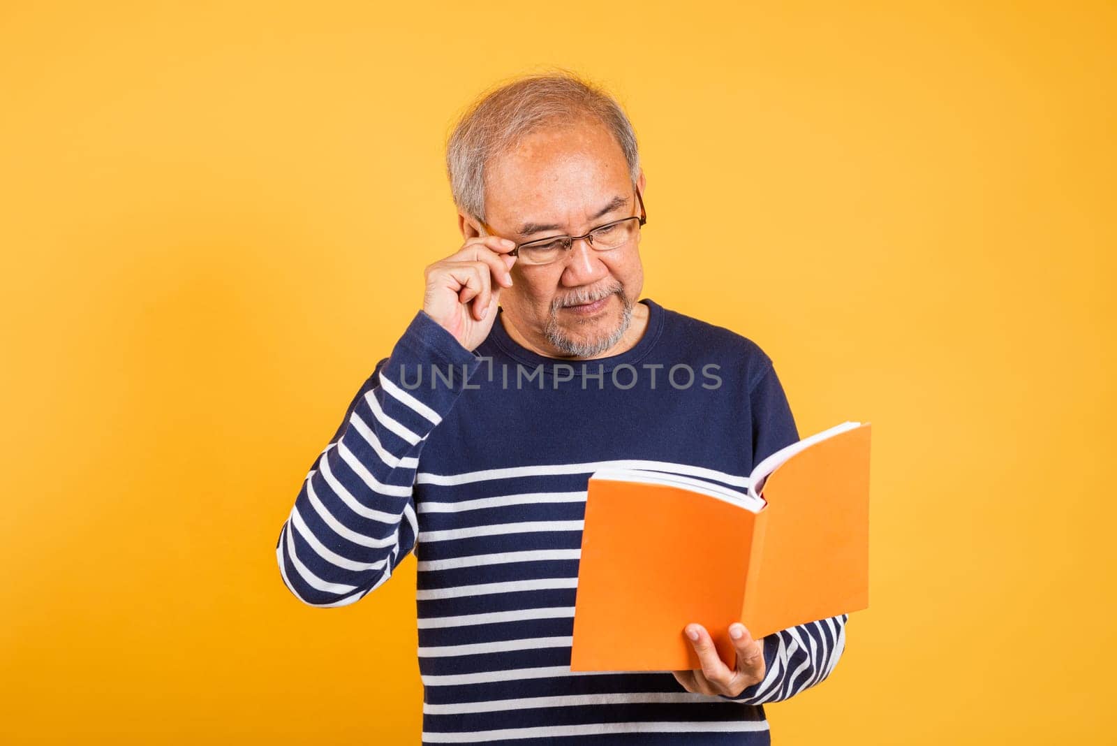 Portrait smiling Asian old man wearing glasses reading a book studio shot isolated yellow background, happy elderly man enjoying interesting story, time to relaxation